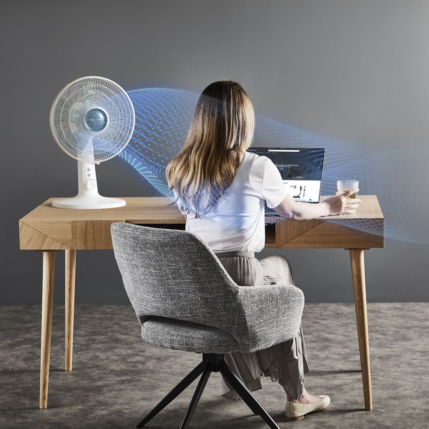 Woman using laptop at desk. A fan blows air towards her. Glass of water on the desk.
