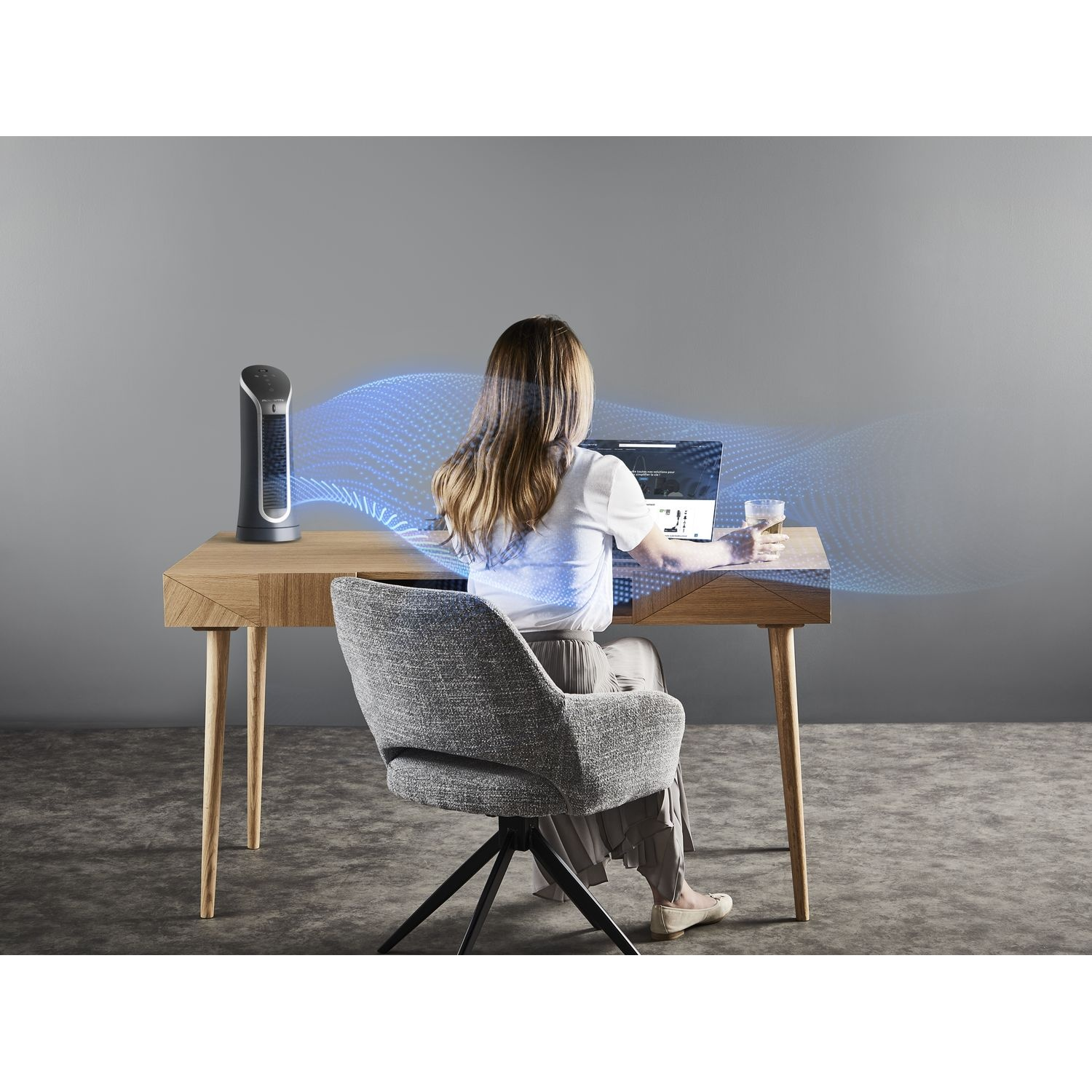 Femme assise à un bureau en bois, utilisant un ordinateur portable, avec un ventilateur émettant de l'air bleu.