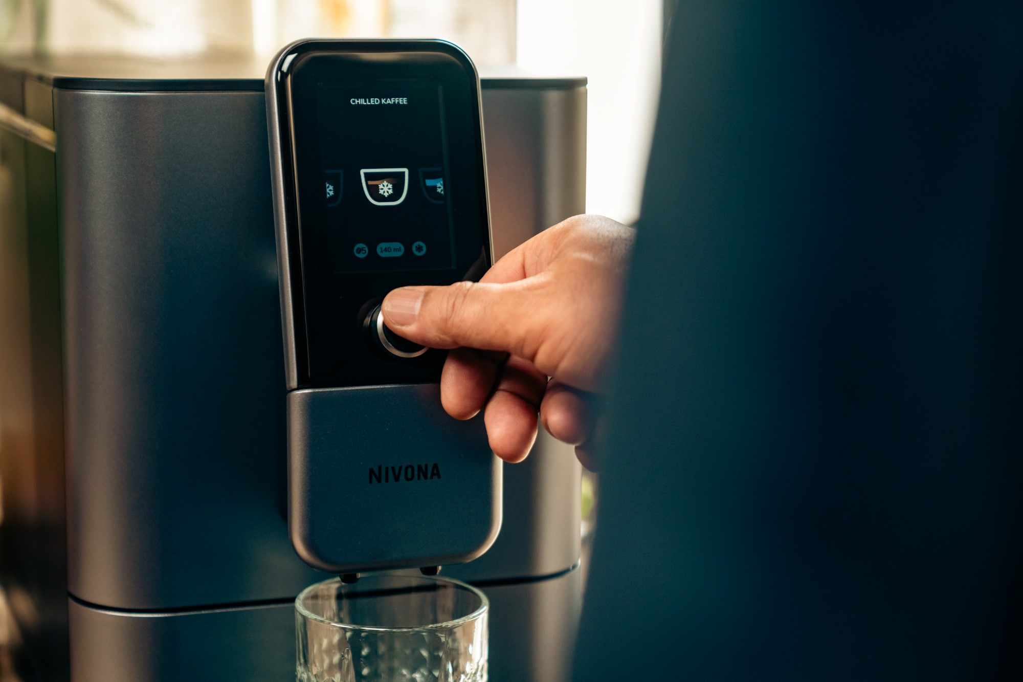 Dłoń dotyka czarnego ekranu na maszynie z logo Nivona. Szklanka z przodu.