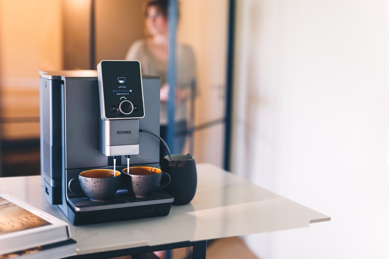 Ekspres do kawy z dwoma filiżankami, na białym stole, rozmyte tło z osobą.