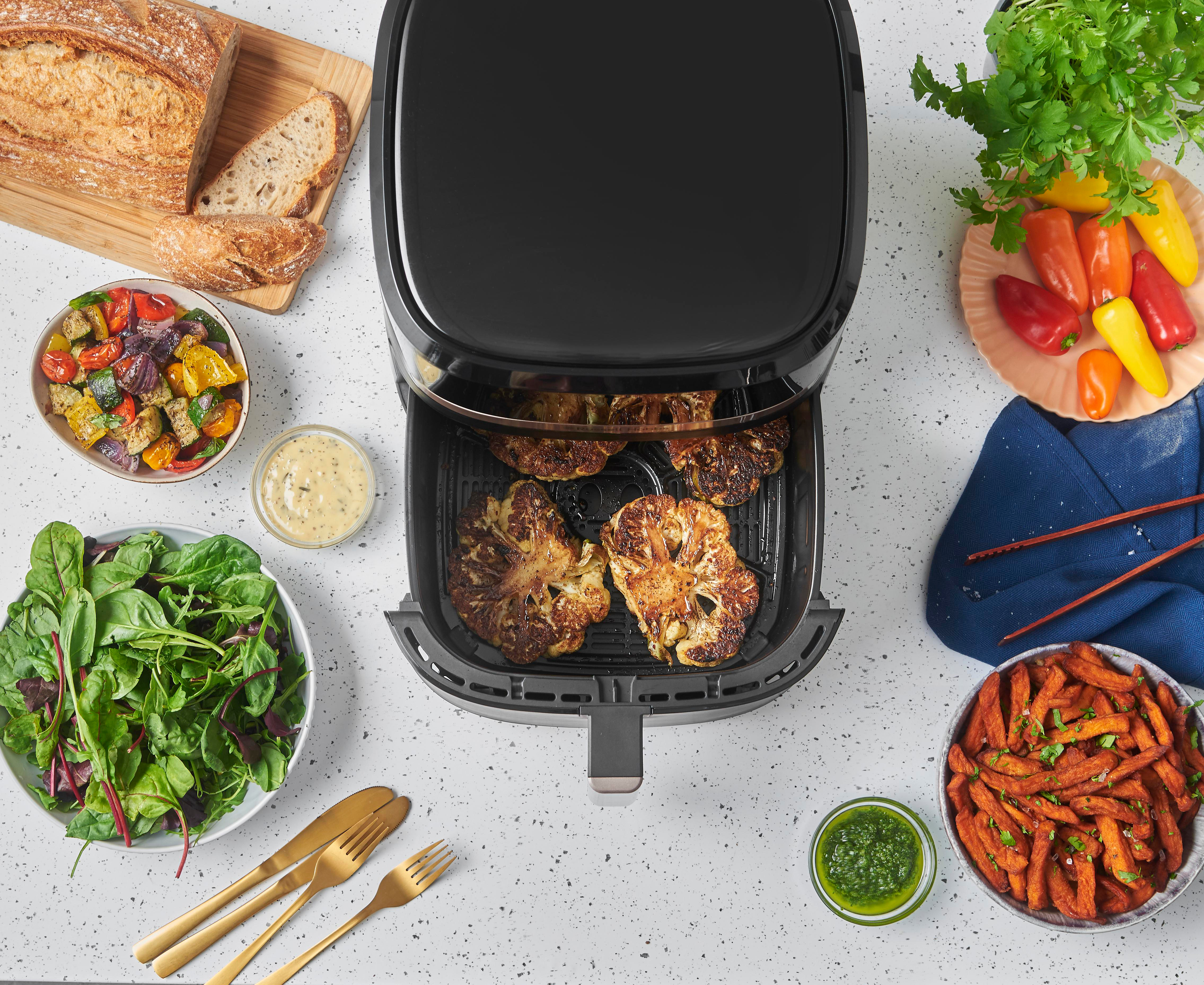 Ein Airfryer mit gekochtem Blumenkohl, Gemüse und anderen Gerichten auf einer weißen Arbeitsplatte.