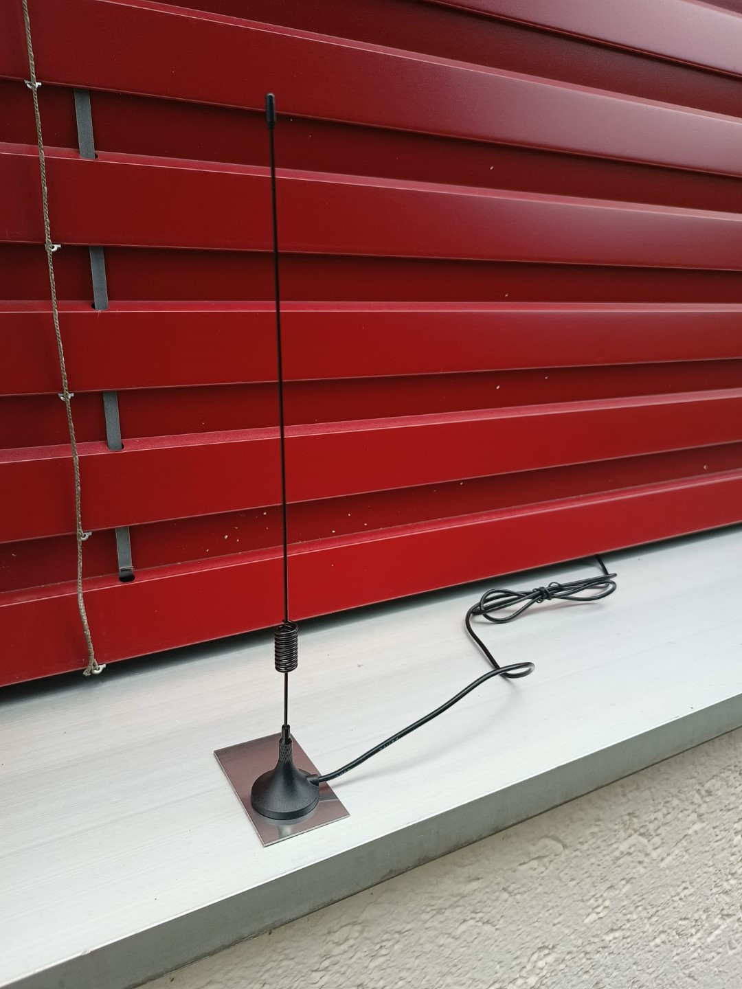 Antenna nera montata su un davanzale. Tende rosse sullo sfondo.