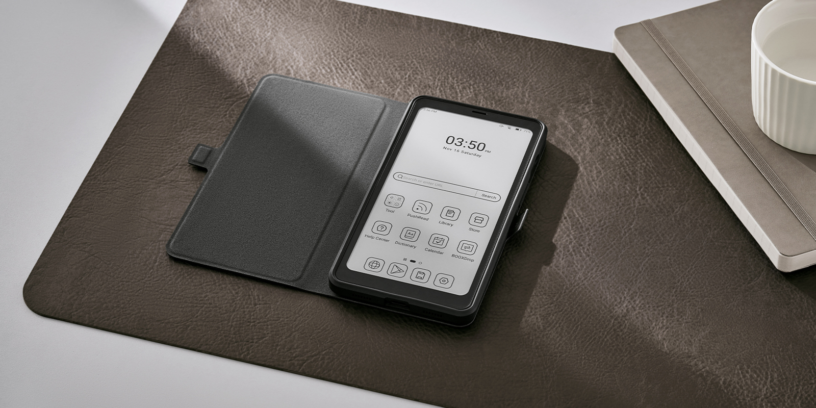 Ein schwarzes Smartphone in einer Hülle, auf einer braunen Matte, mit Notizbuch und Tasse.