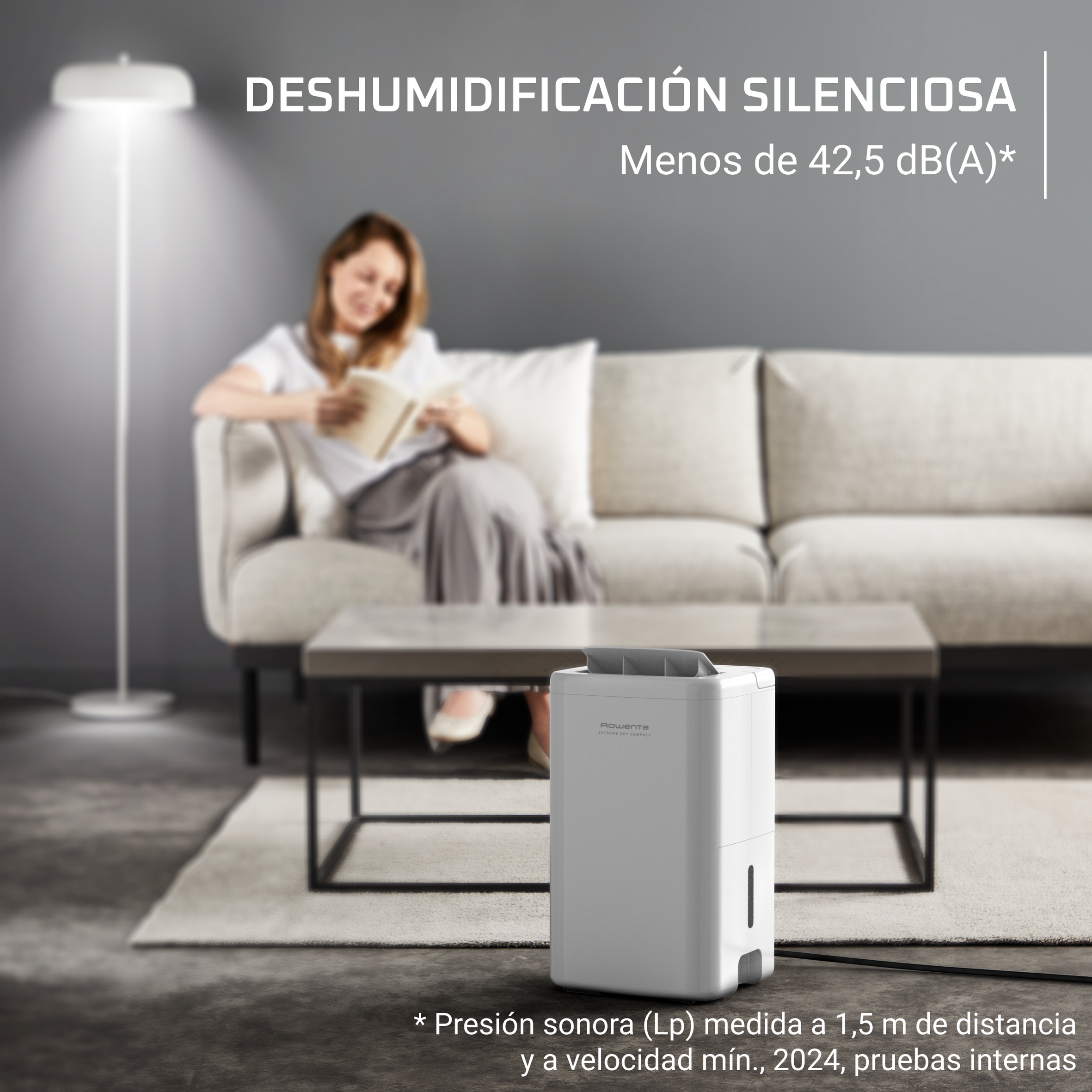 Una mujer leyendo en un sofá con un deshumidificador blanco en el suelo. Una lámpara de pie ilumina la escena.