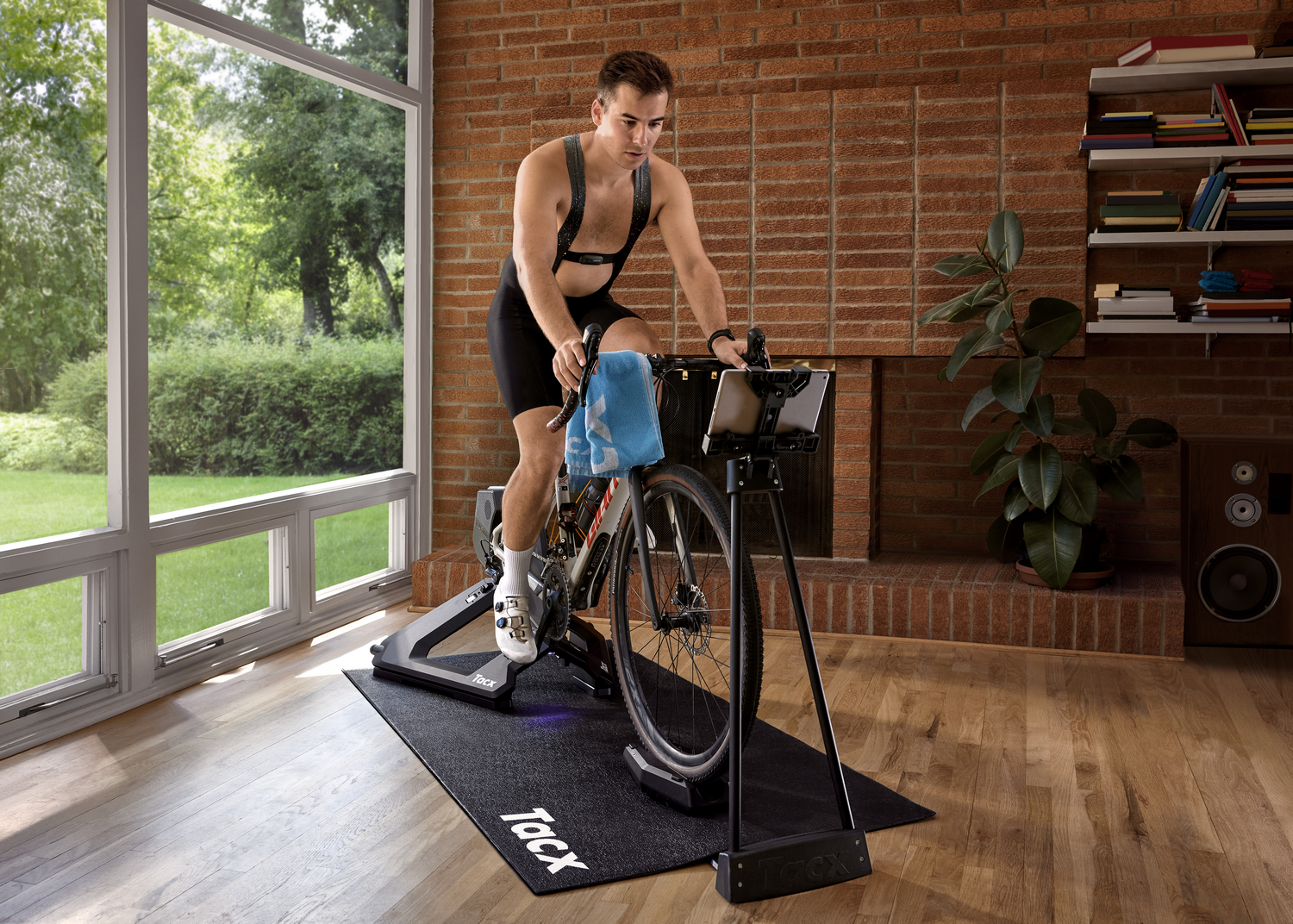 Hombre en bicicleta estática. Lleva un monitor de frecuencia cardíaca y una toalla en el manillar. Habitación con ventana y pared de ladrillo.