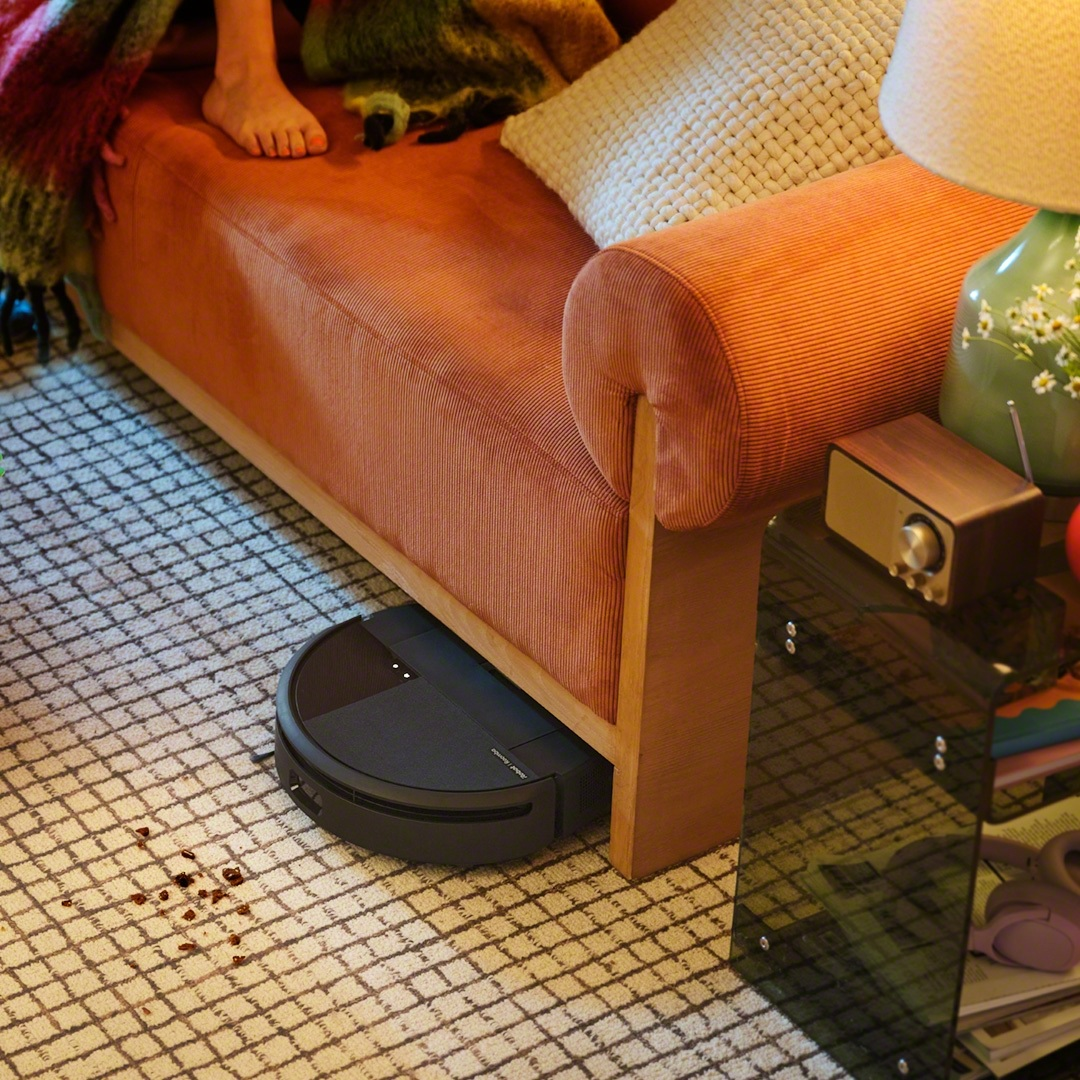 Robot sprzątający pod sofą. Widoczne stopy osoby. Dywan i meble w zasięgu wzroku.