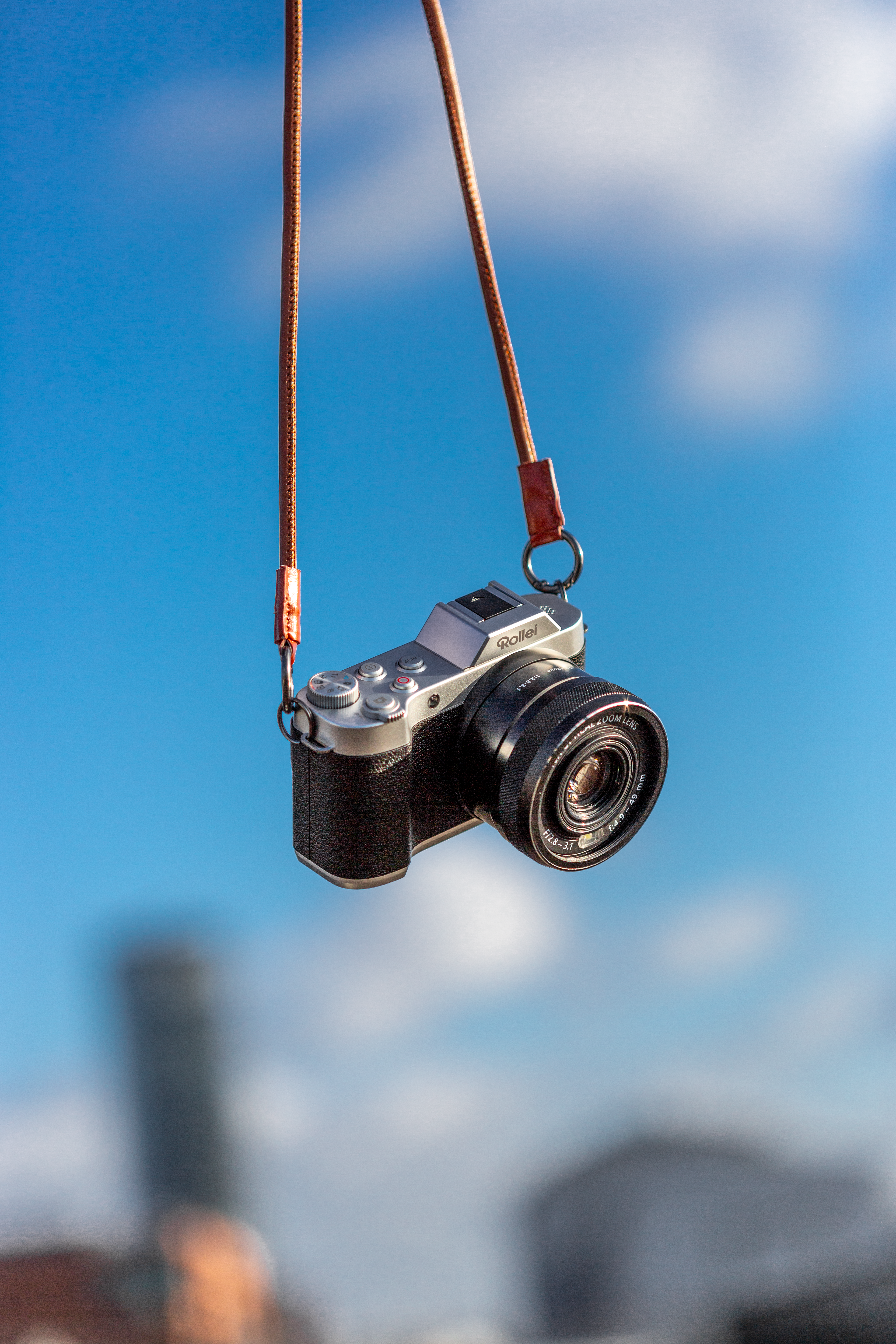 Una fotocamera Rollei è appesa a un cinturino marrone contro un cielo blu con edifici sfocati.