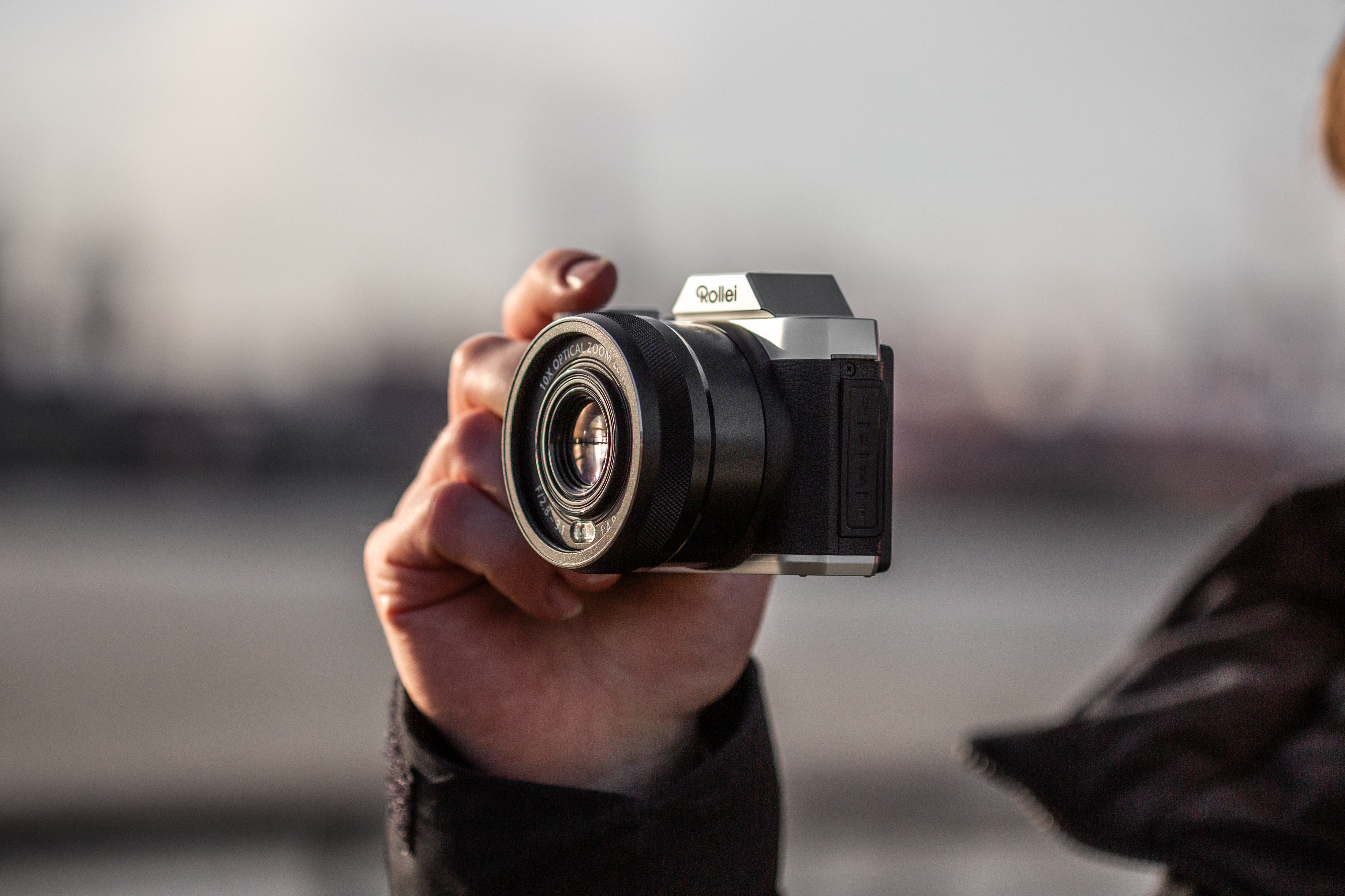 Una persona che tiene in mano una fotocamera Rollei nera e argento, sfondo sfocato di una città.
