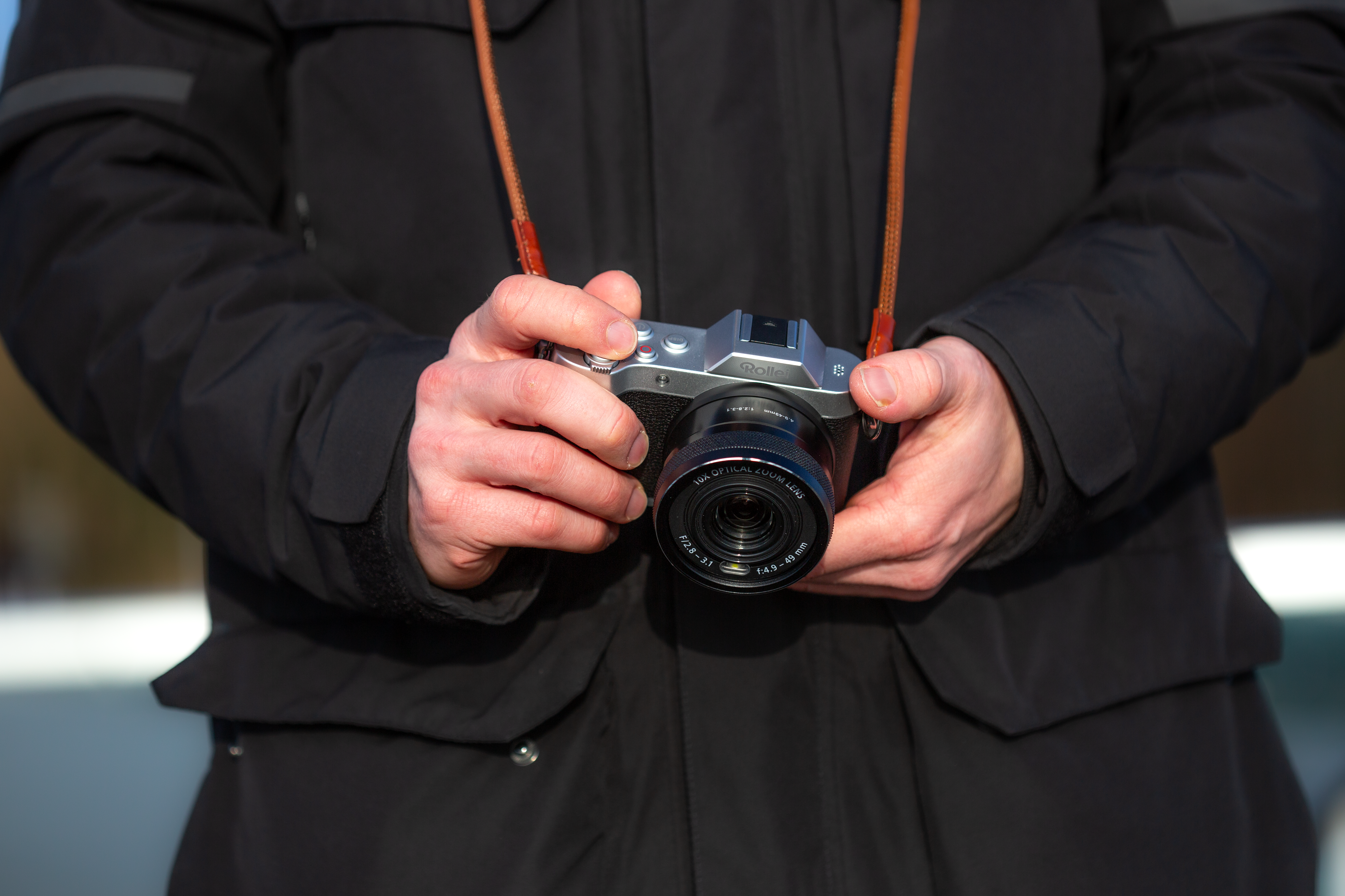Una persona che tiene una fotocamera argentata con cinturino marrone, cappotto invernale.