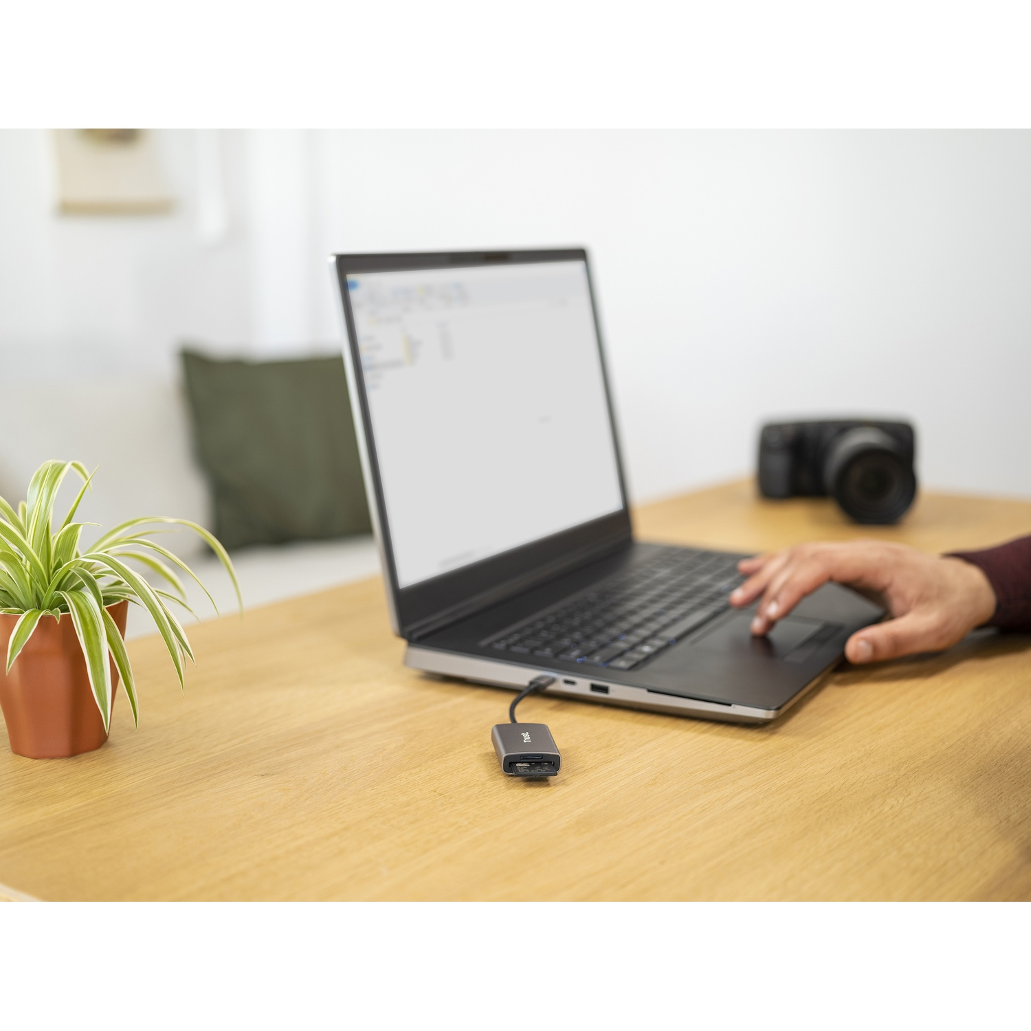Un laptop con un adattatore USB su una scrivania di legno. Sono visibili anche una pianta in vaso e una fotocamera.