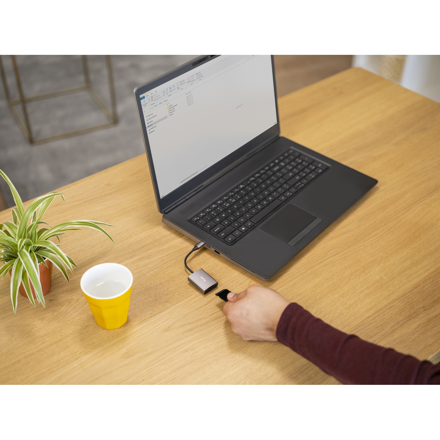 Laptop con hub USB-C e scheda di memoria. Una tazza gialla e una piccola pianta sul tavolo.