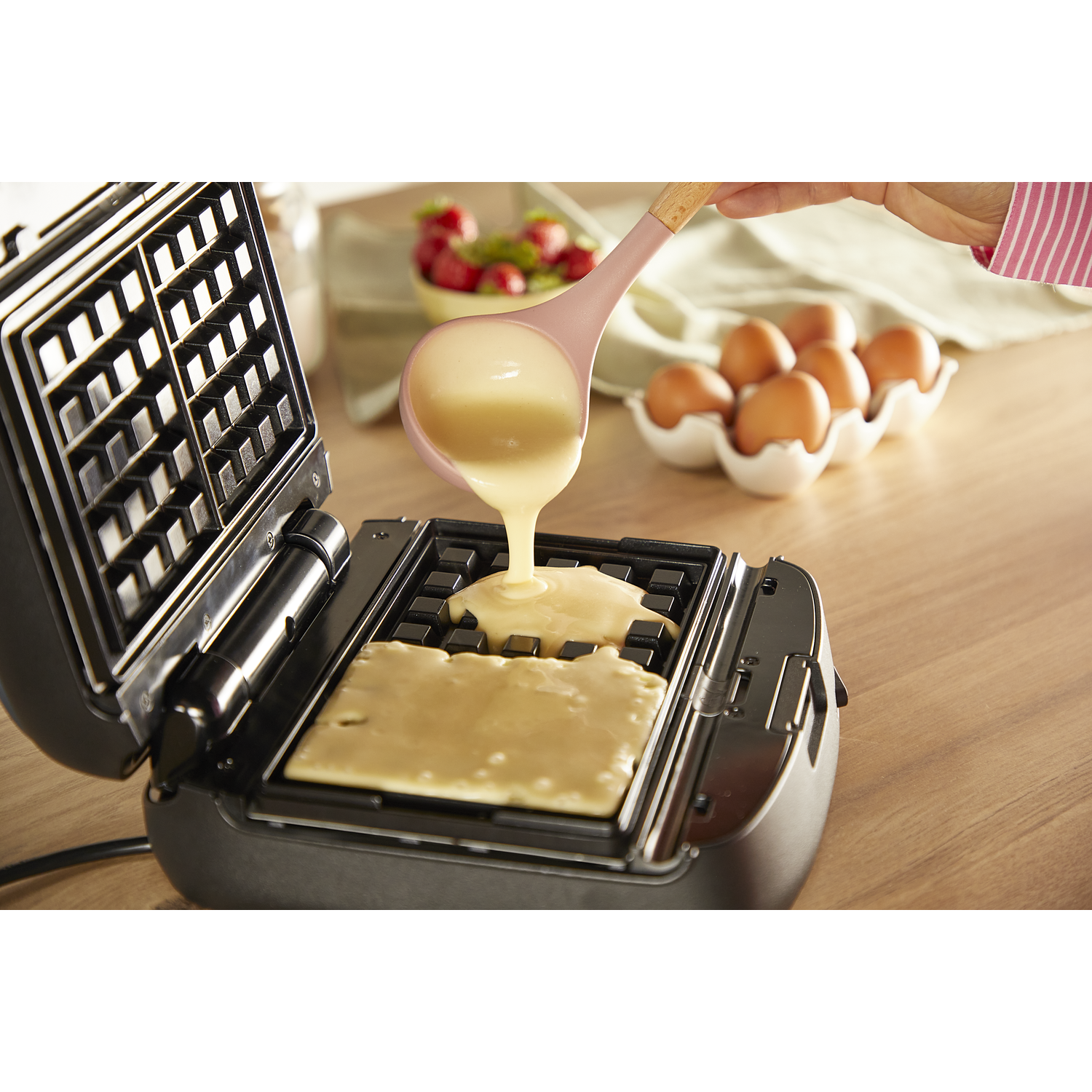 Gaufrier avec pâte versée, fraises et œufs en arrière-plan sur une table.