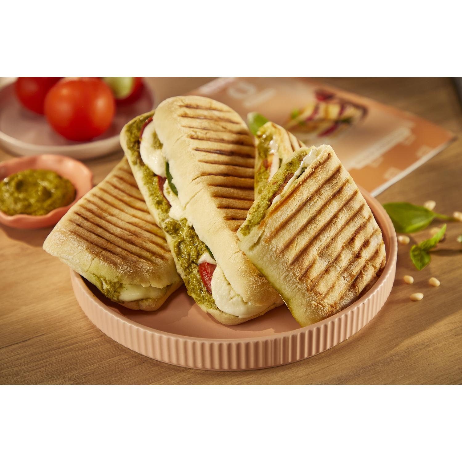 Trois sandwichs panini grillés au pesto, mozzarella et tomate sur une assiette rose. Tomates à l'arrière.