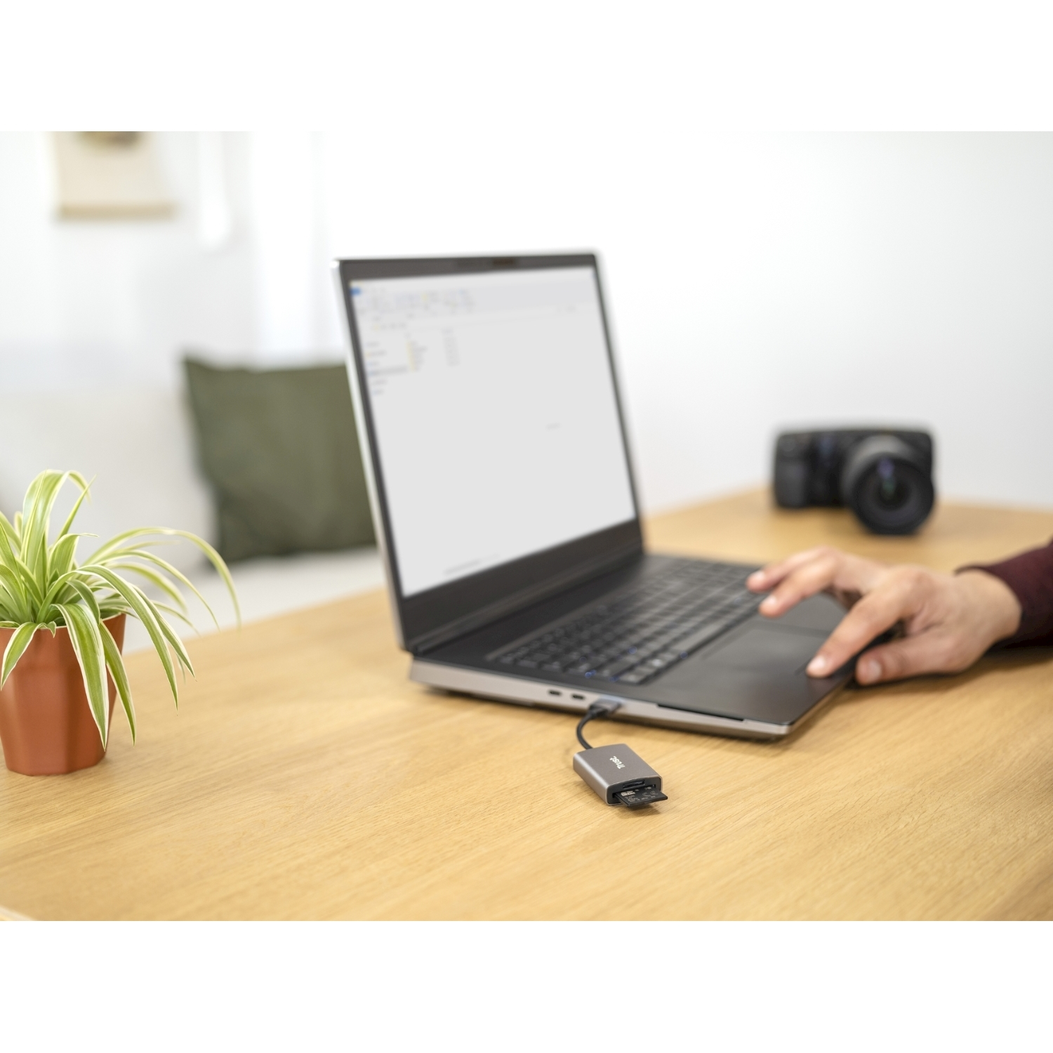Laptop con adattatore USB, su scrivania in legno con pianta e fotocamera.