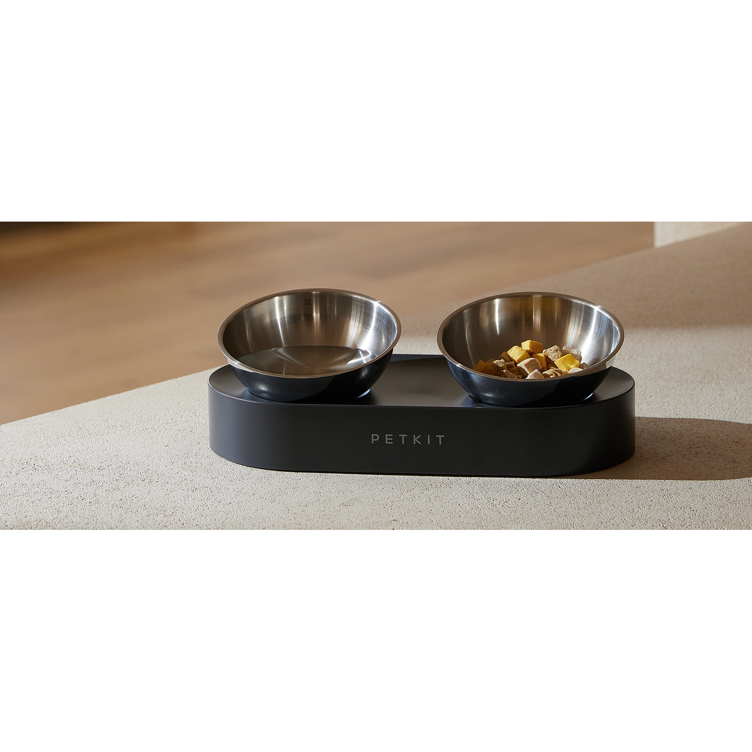 Two stainless steel pet food bowls on a black base, placed on a countertop.