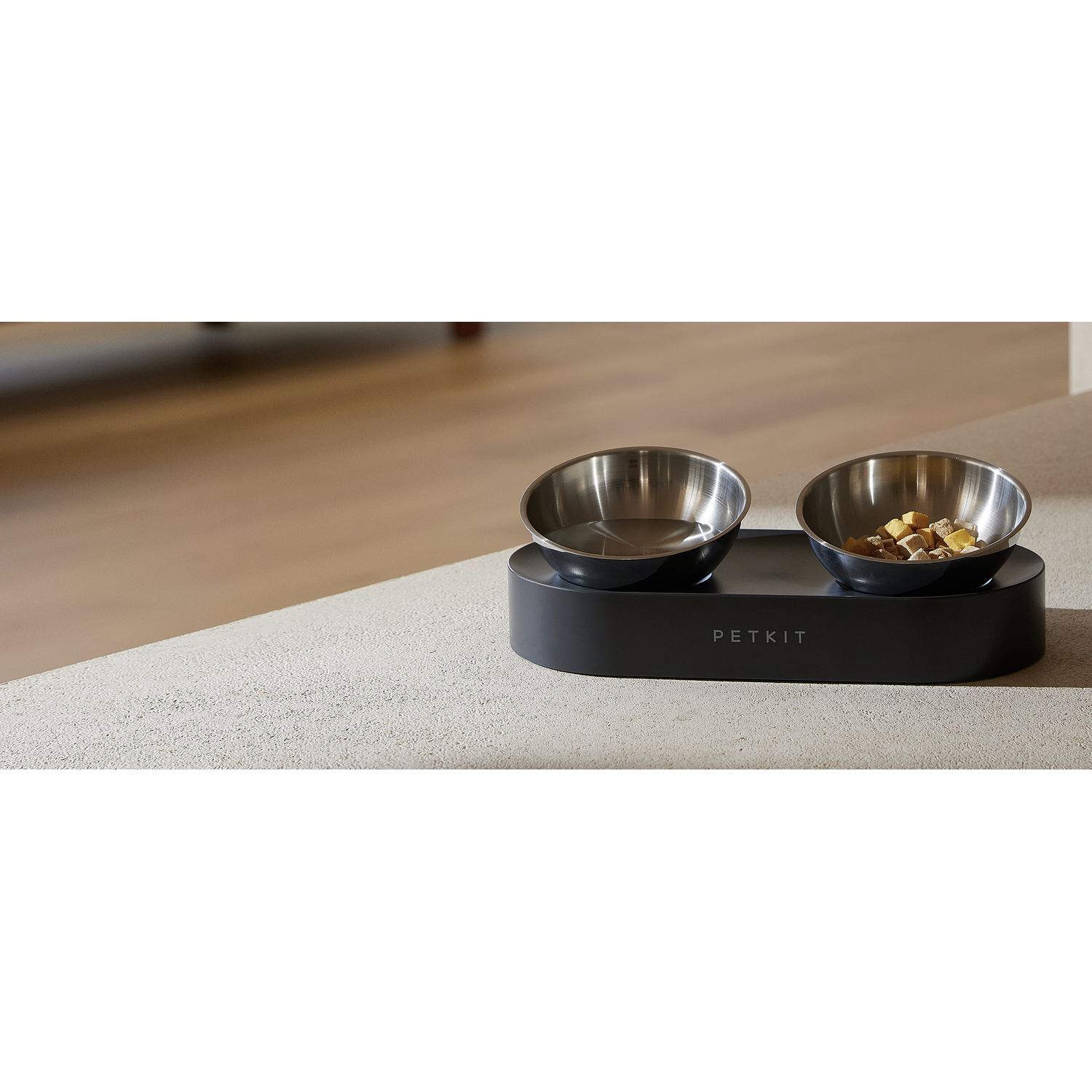 Two stainless steel pet bowls on a dark gray base, with food in one. Background is a neutral surface.