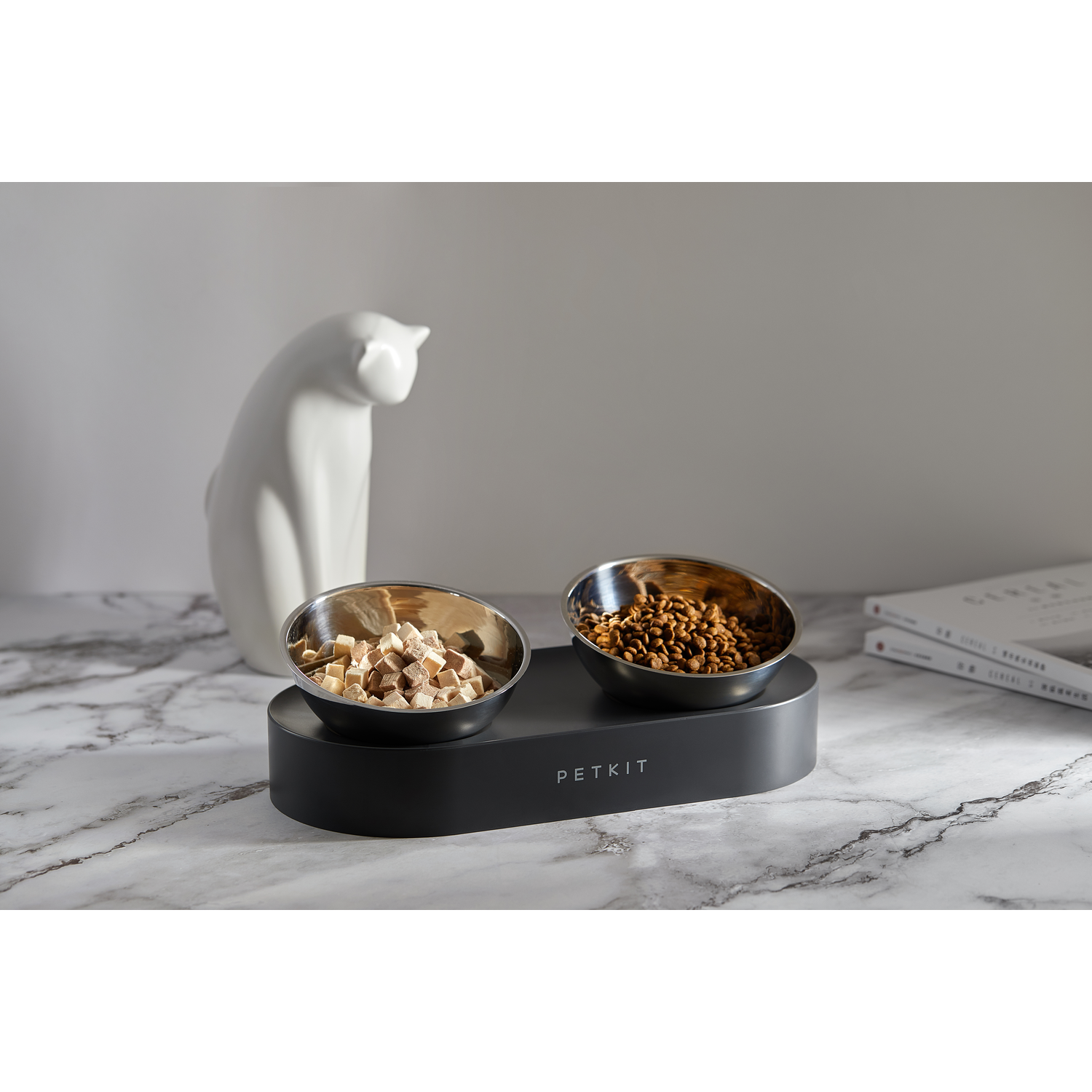Pet food in stainless steel bowls on a dark gray base, with a white cat statue.