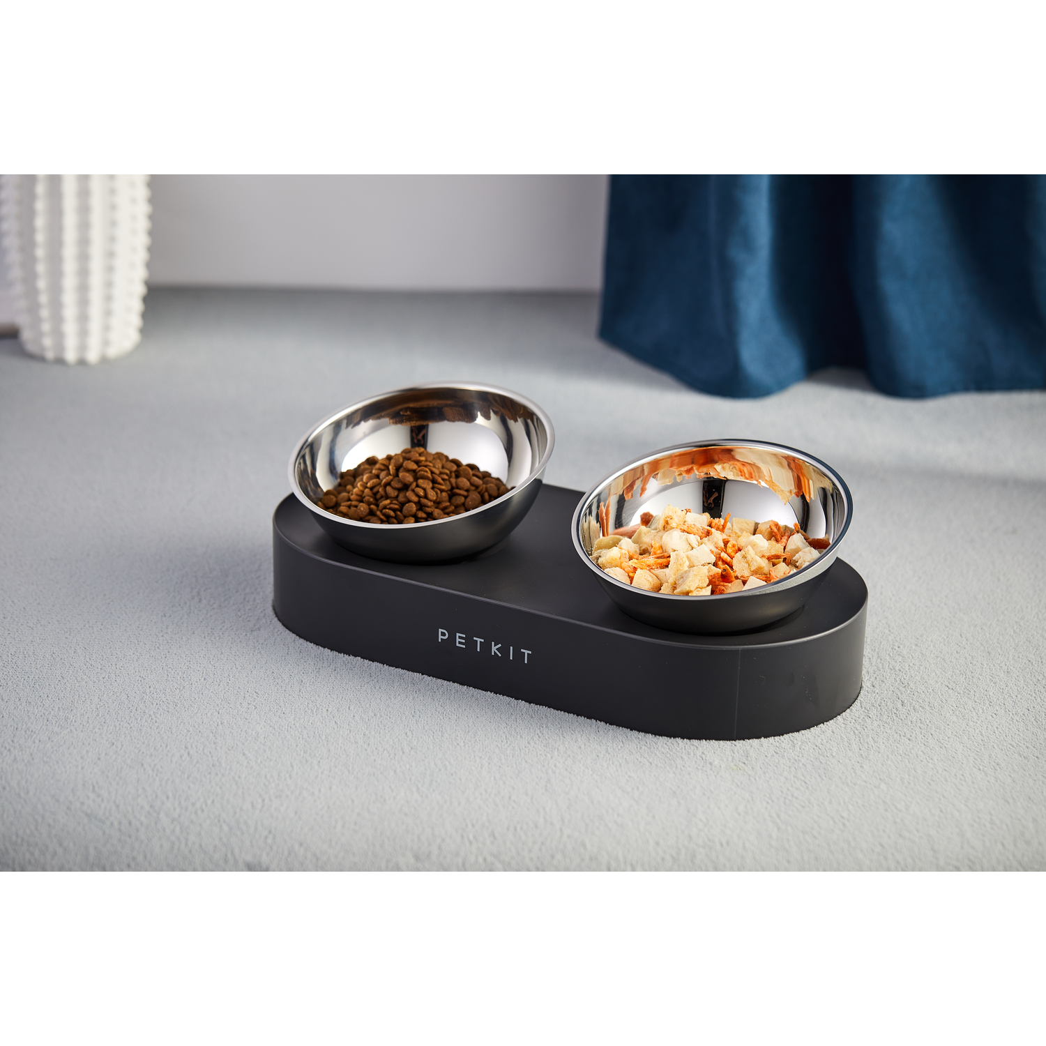 A black pet bowl set with two stainless steel bowls, one with brown kibble and the other with food pieces.