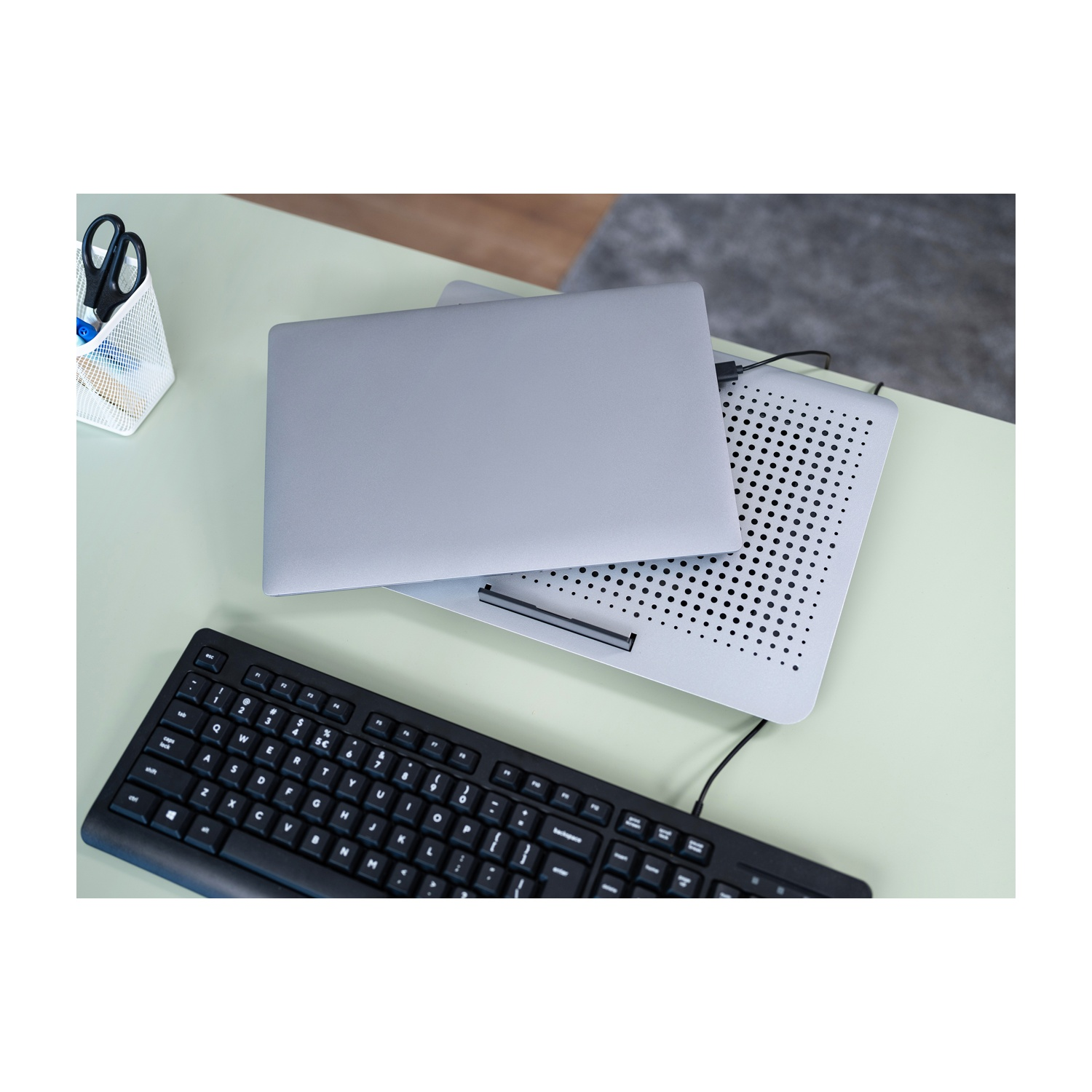 Un laptop grigio su un supporto di raffreddamento argentato, tastiera nera davanti. Tutto su una scrivania verde chiaro.