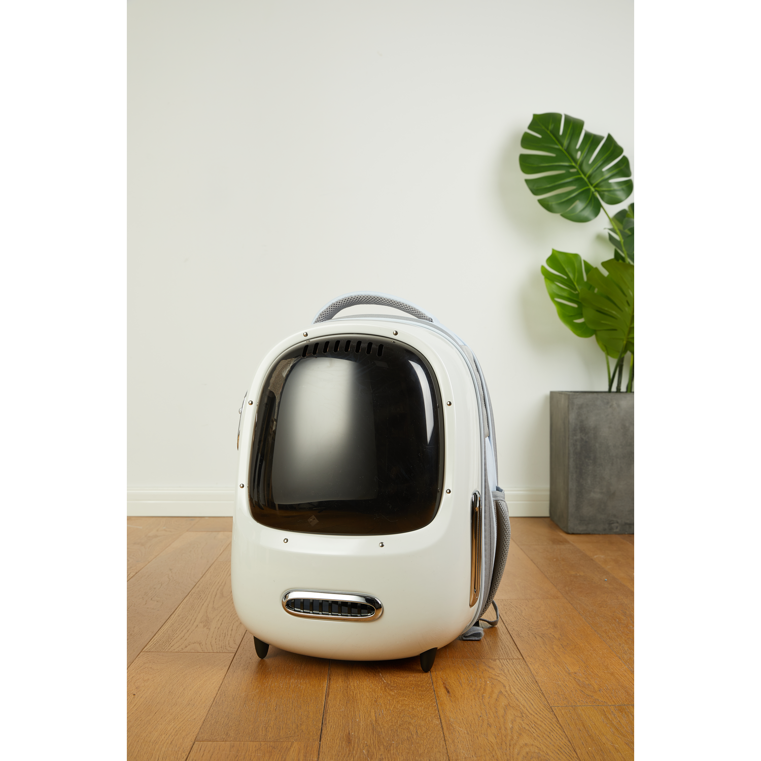 White pet carrier backpack. It has a clear front window, sitting on wooden floor. A plant in background.