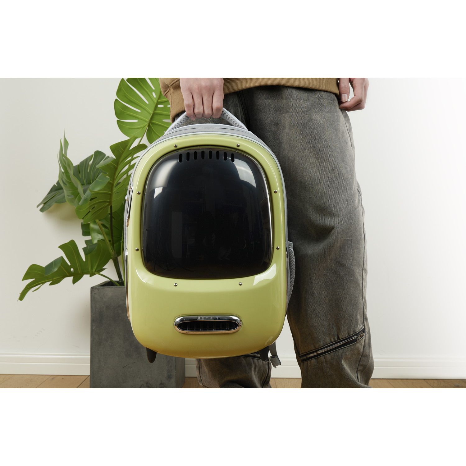 A person holds a green pet carrier with a clear front panel, standing by a plant in a pot.