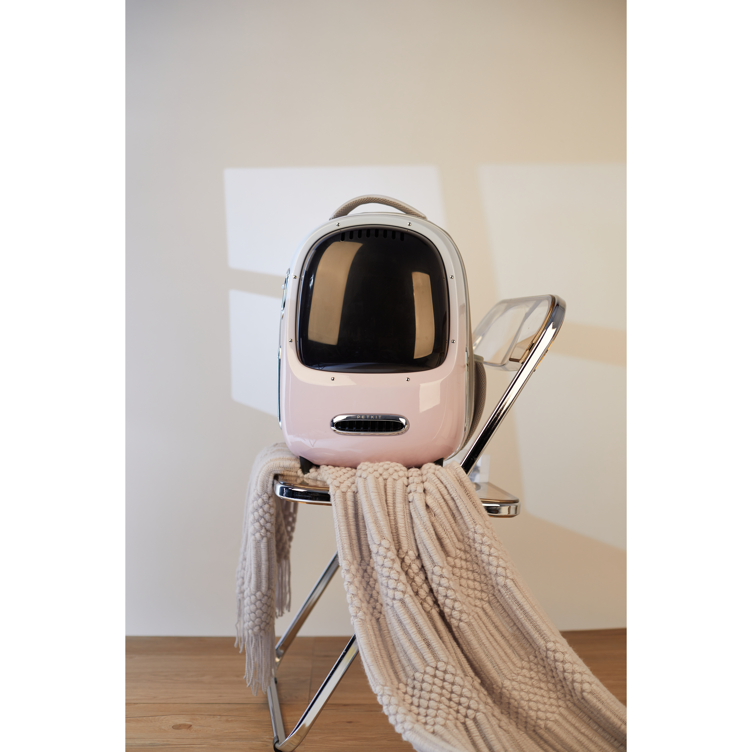 A pink and white pet carrier on a chair with a blanket. The carrier has a clear front.