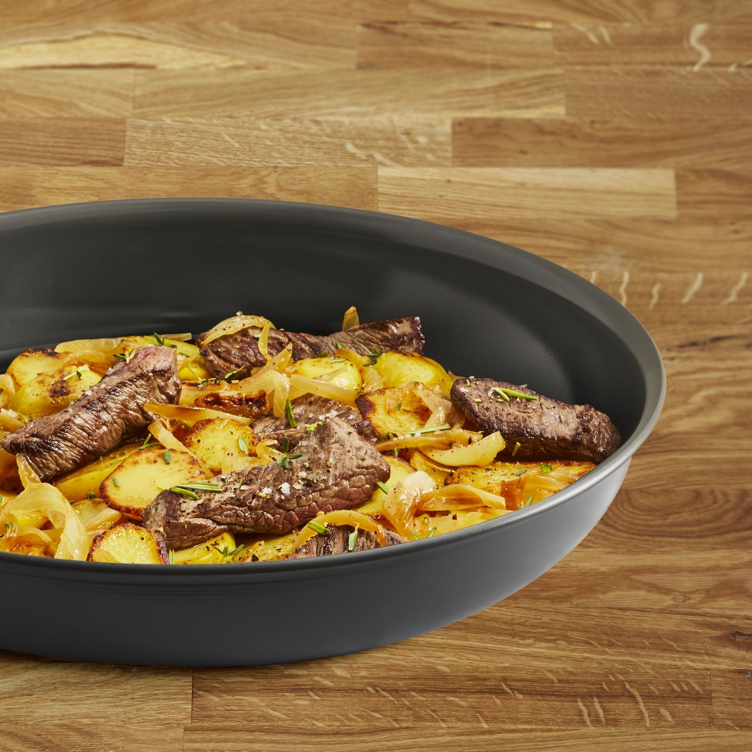 Beef and potatoes in a black pan on a wooden table. The dish includes beef steaks, potatoes, and onions.