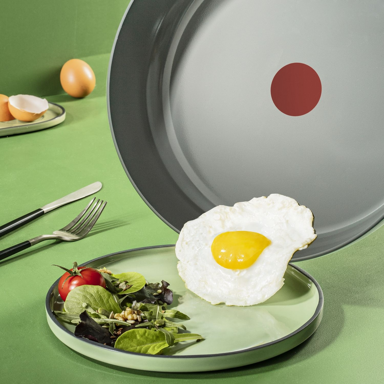 A fried egg with a yellow yolk sits on a green plate with salad. A large pan with a red dot and other eggs are in the background.