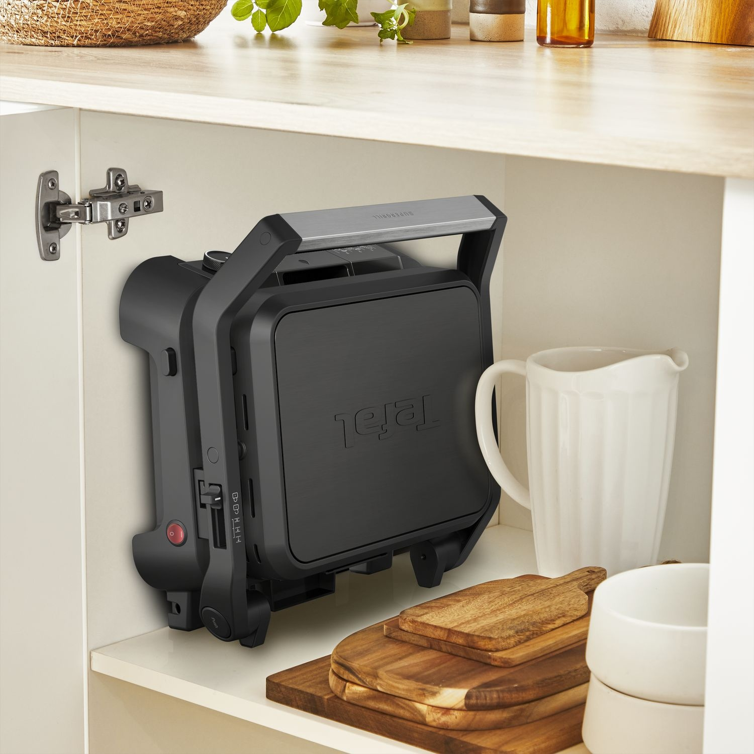 A black panini press, stored inside a white cabinet with a pitcher and wooden boards.