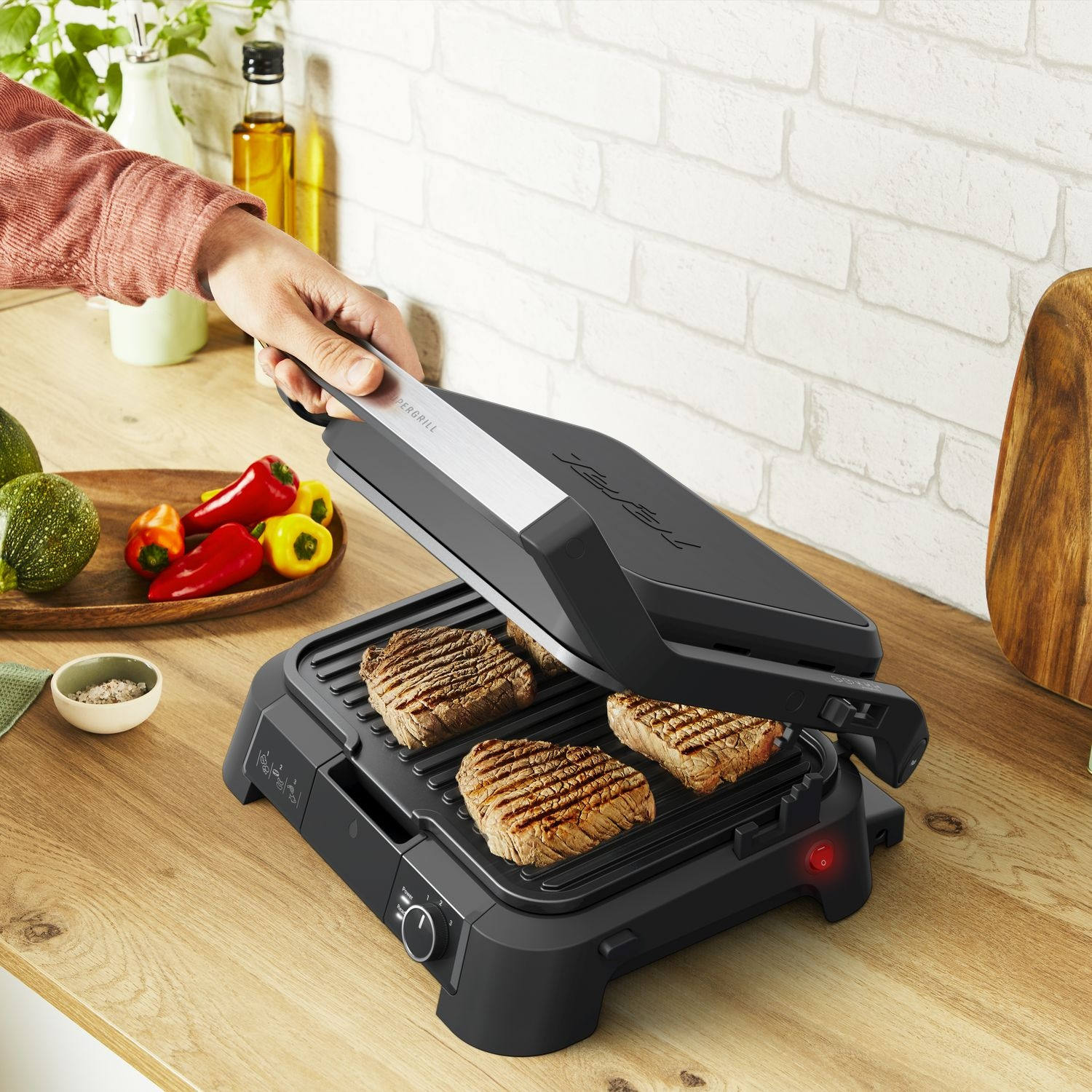 A person grilling steaks on a black electric grill, with vegetables on the side.