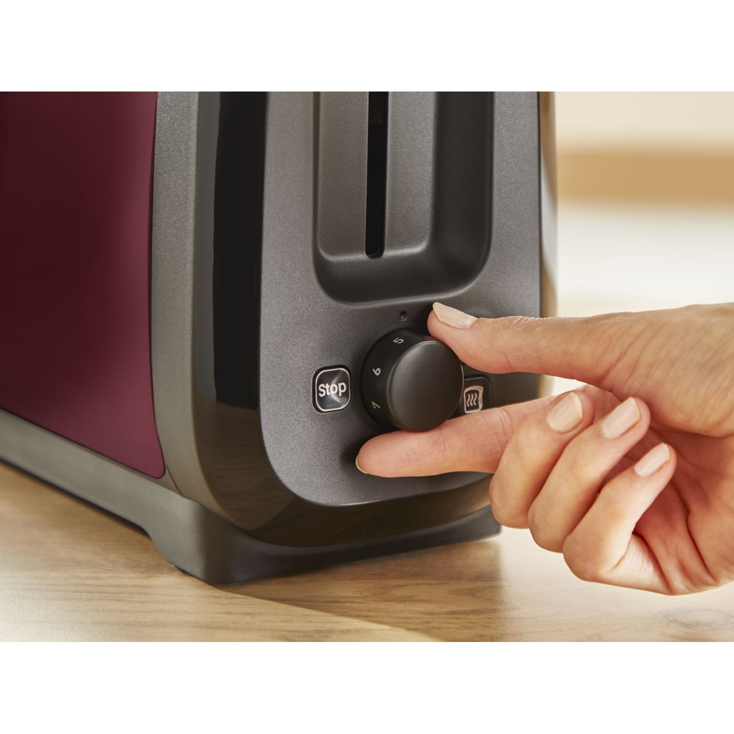 A hand turning the knob of a burgundy toaster on a wooden surface, with a stop button.
