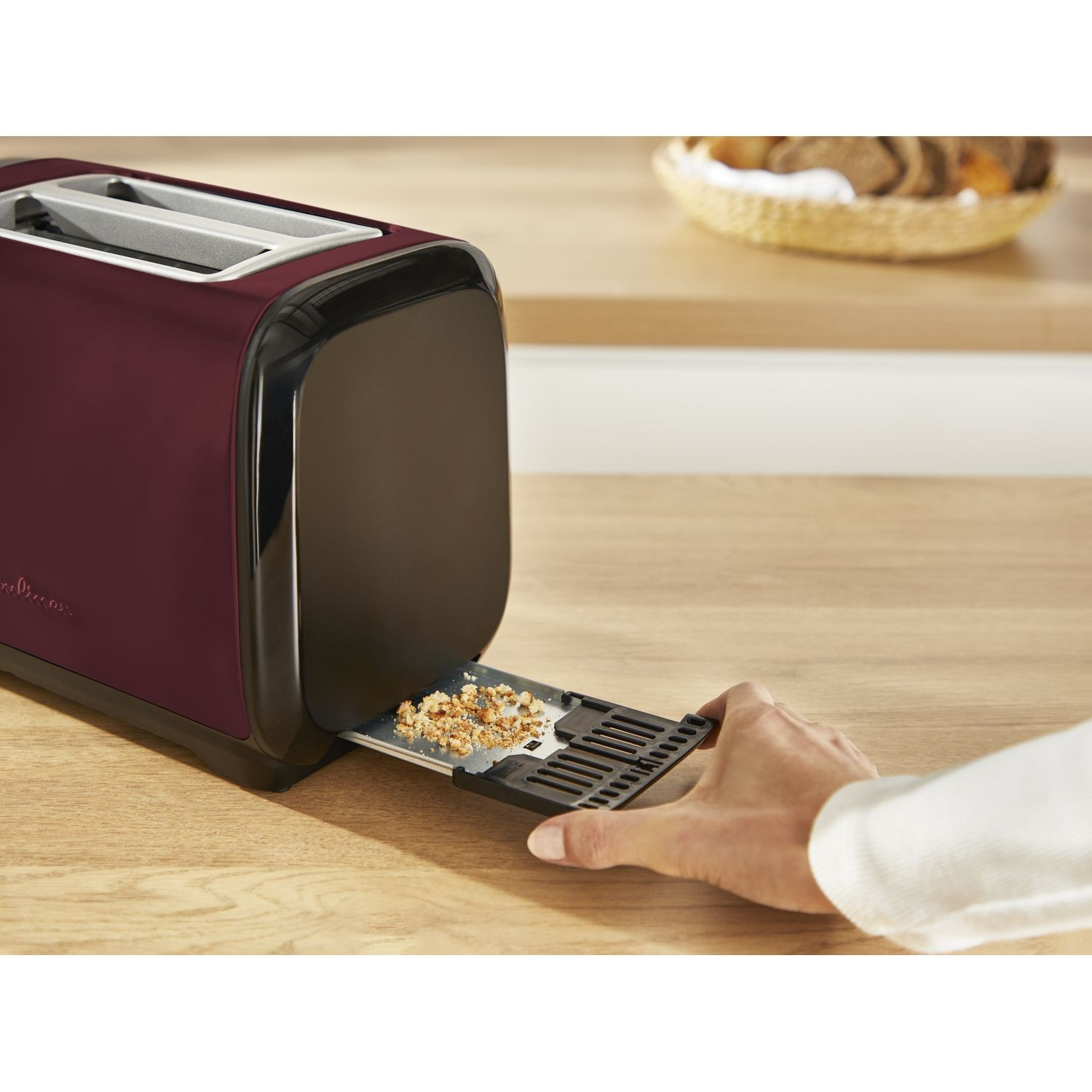 A maroon colored toaster with a hand pulling out a tray with crumbs, on a wooden table.