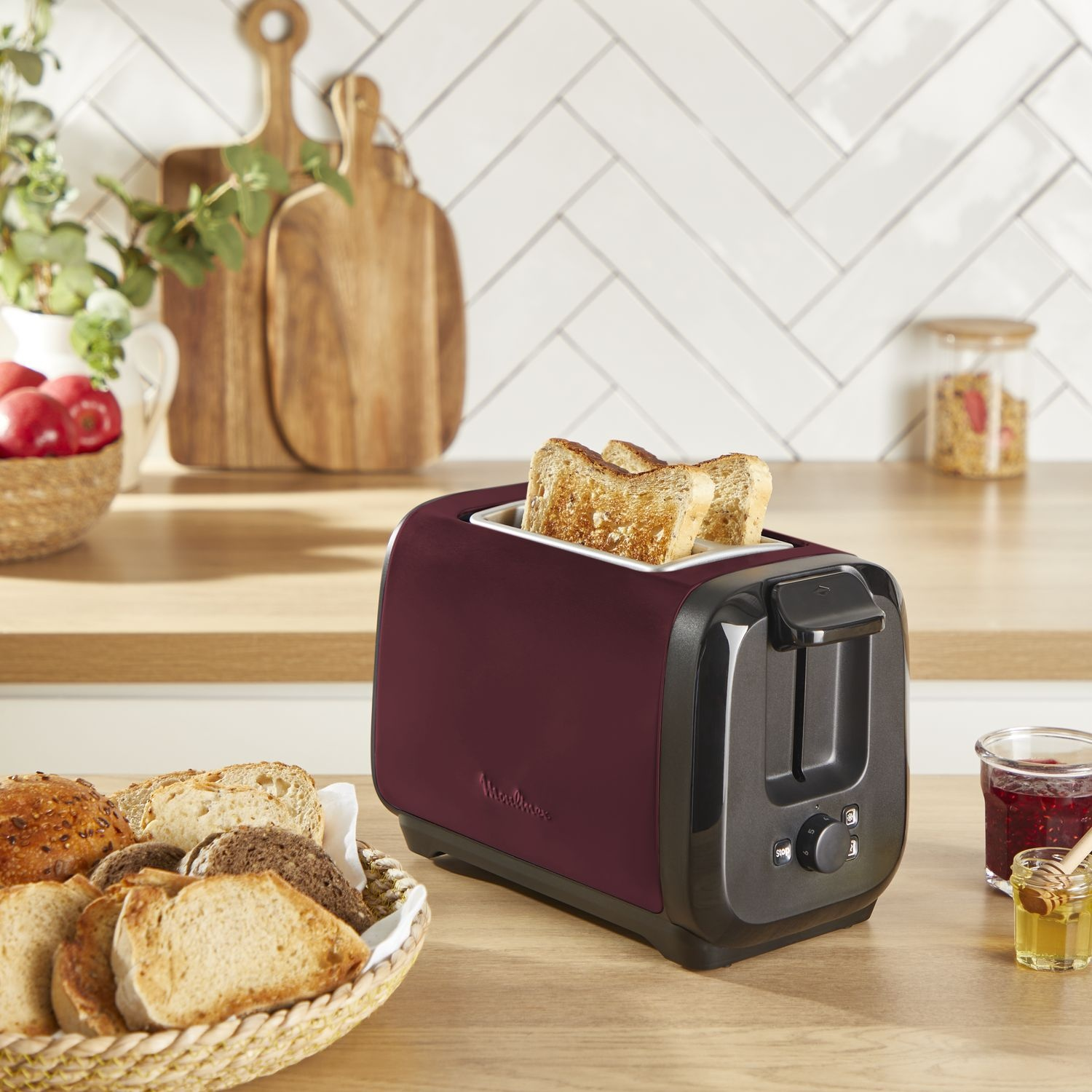 Toaster with bread slices in it, surrounded by bread, jam, and honey on a kitchen counter.