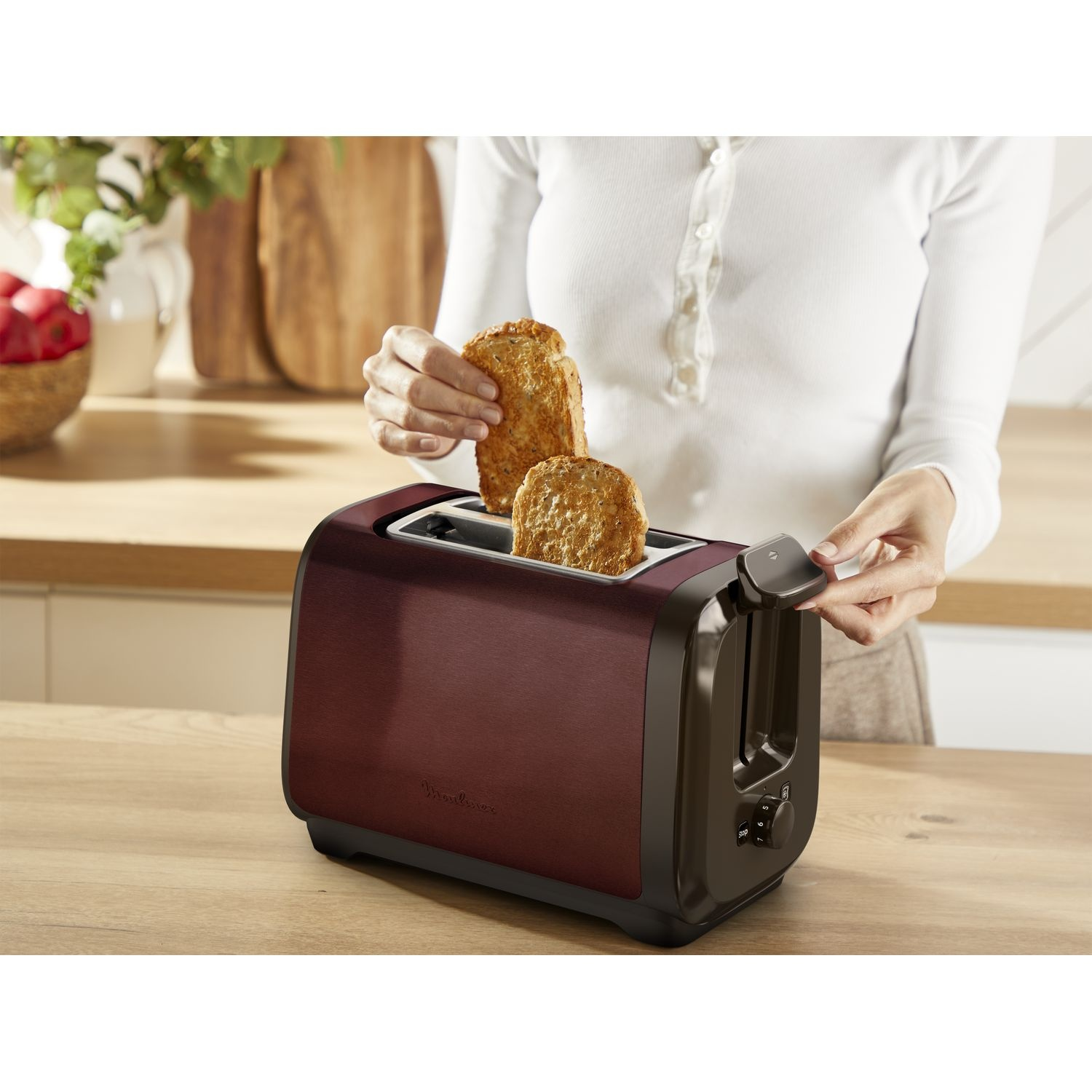 A person taking toast out of a dark red toaster. Wooden table in the foreground.