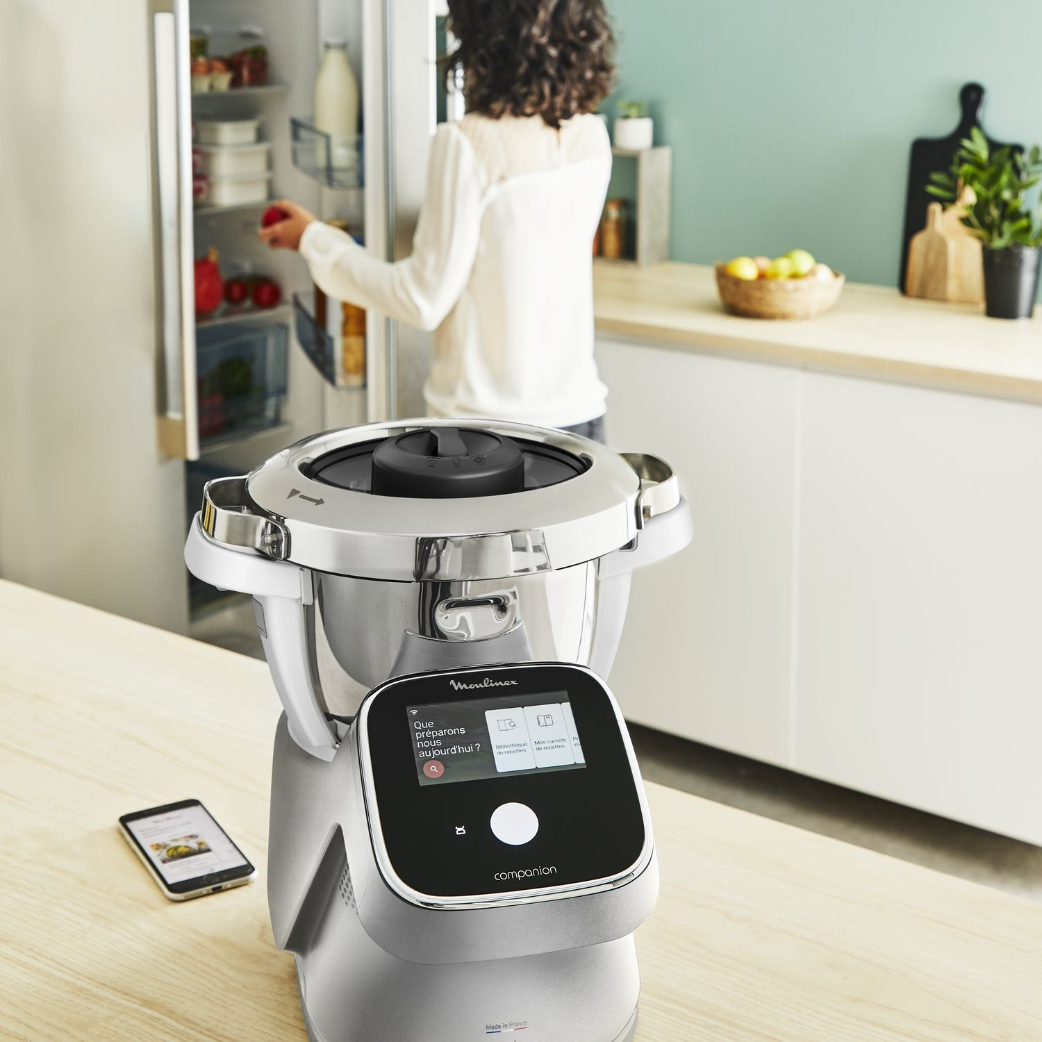 A kitchen appliance with a woman in the background near a refrigerator.