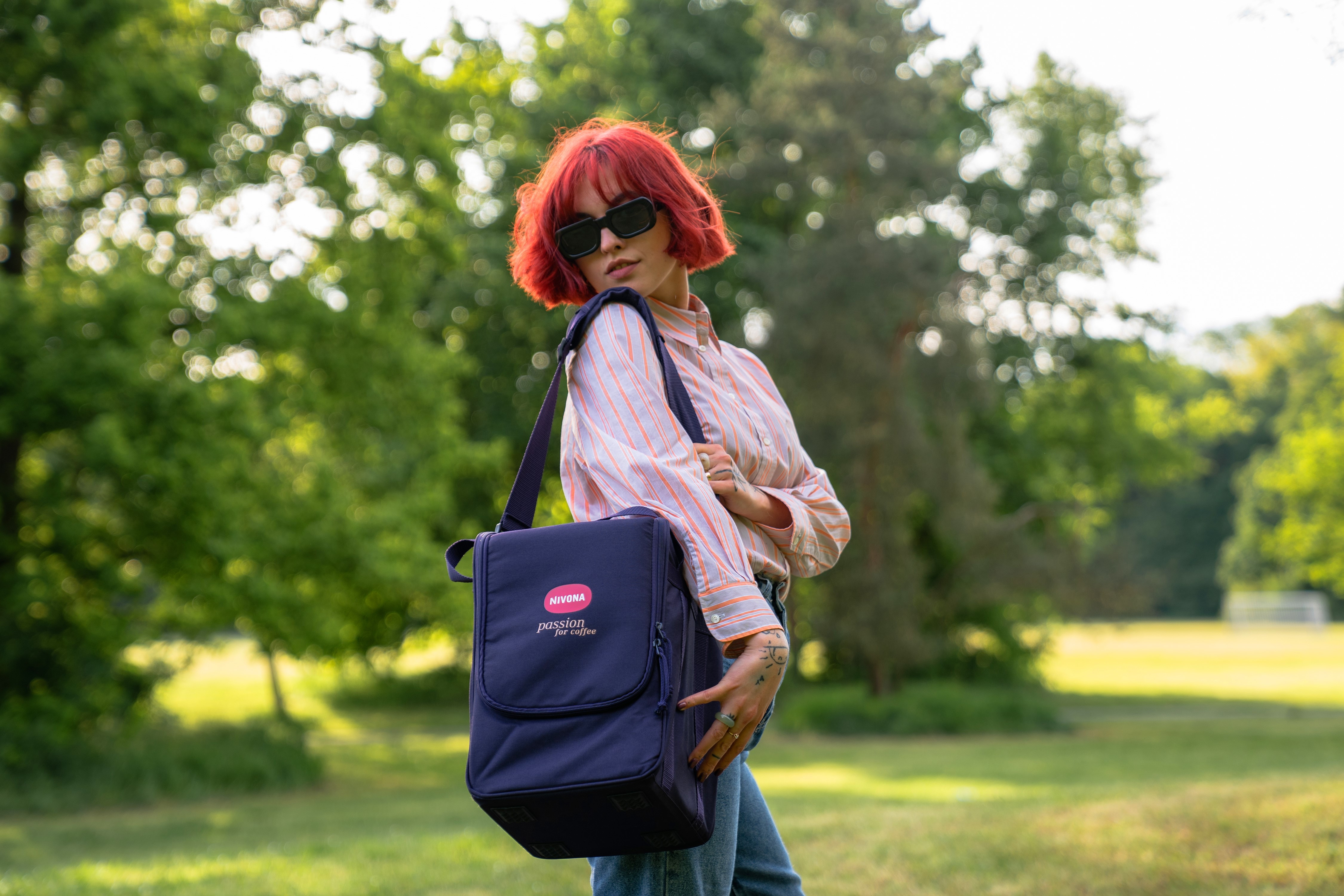 Kobieta z rudymi włosami i okularami przeciwsłonecznymi trzyma niebieską torbę w zielonym parku.