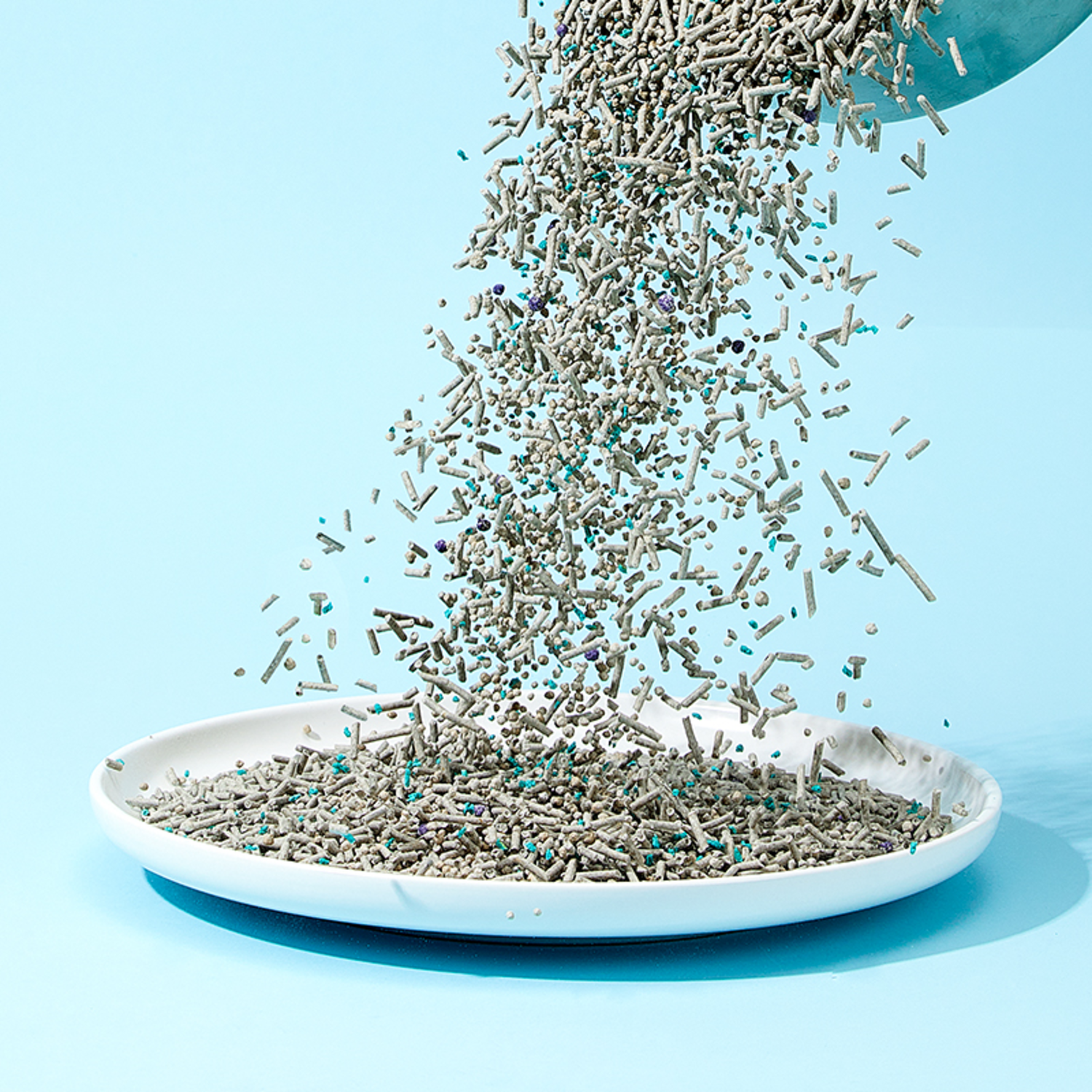 Cat litter pouring into a white dish. The litter is gray-brown with green and blue specks.