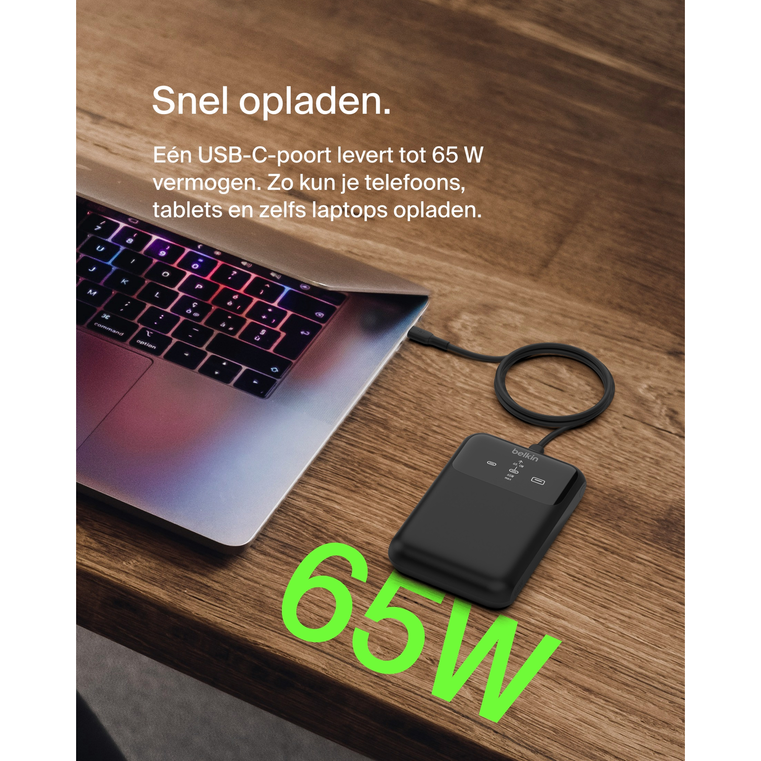 A black Belkin power bank charging a laptop from a USB-C port on a wooden table.