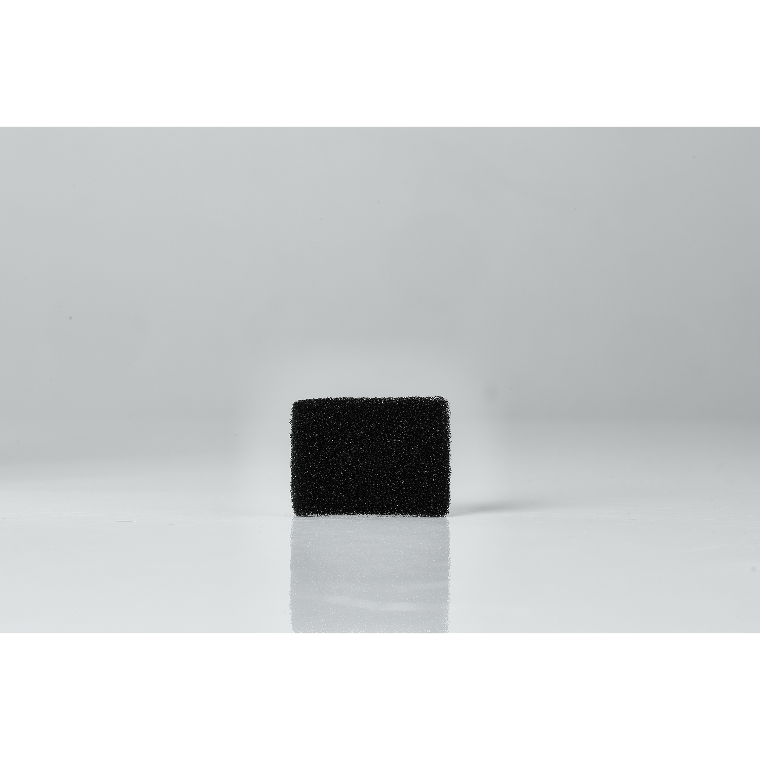 A small, black rectangular foam filter on a white, reflective surface. It has a textured surface.