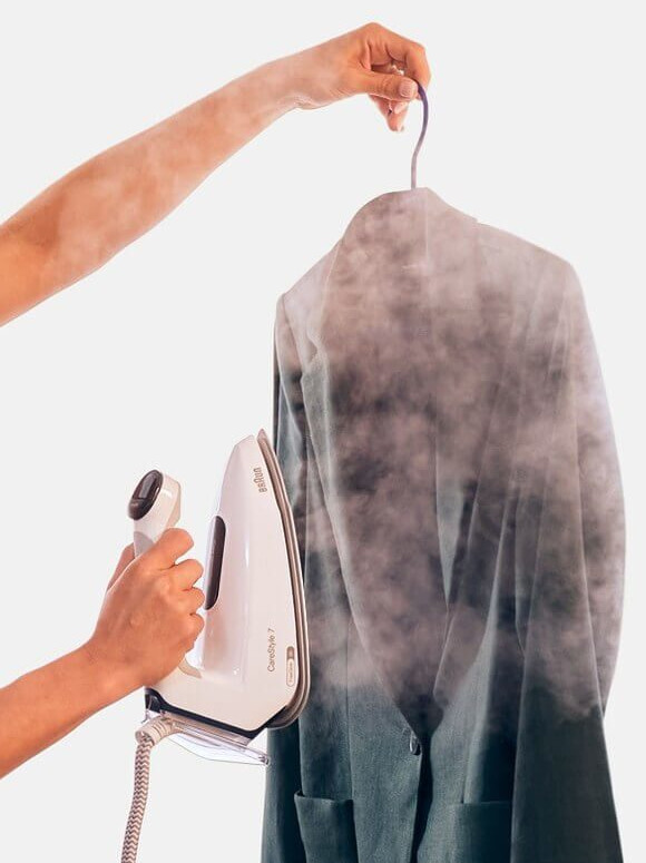 Osoba prasuje turkusową marynarkę białym żelazkiem. Para unosi się z żelazka i kurtki.