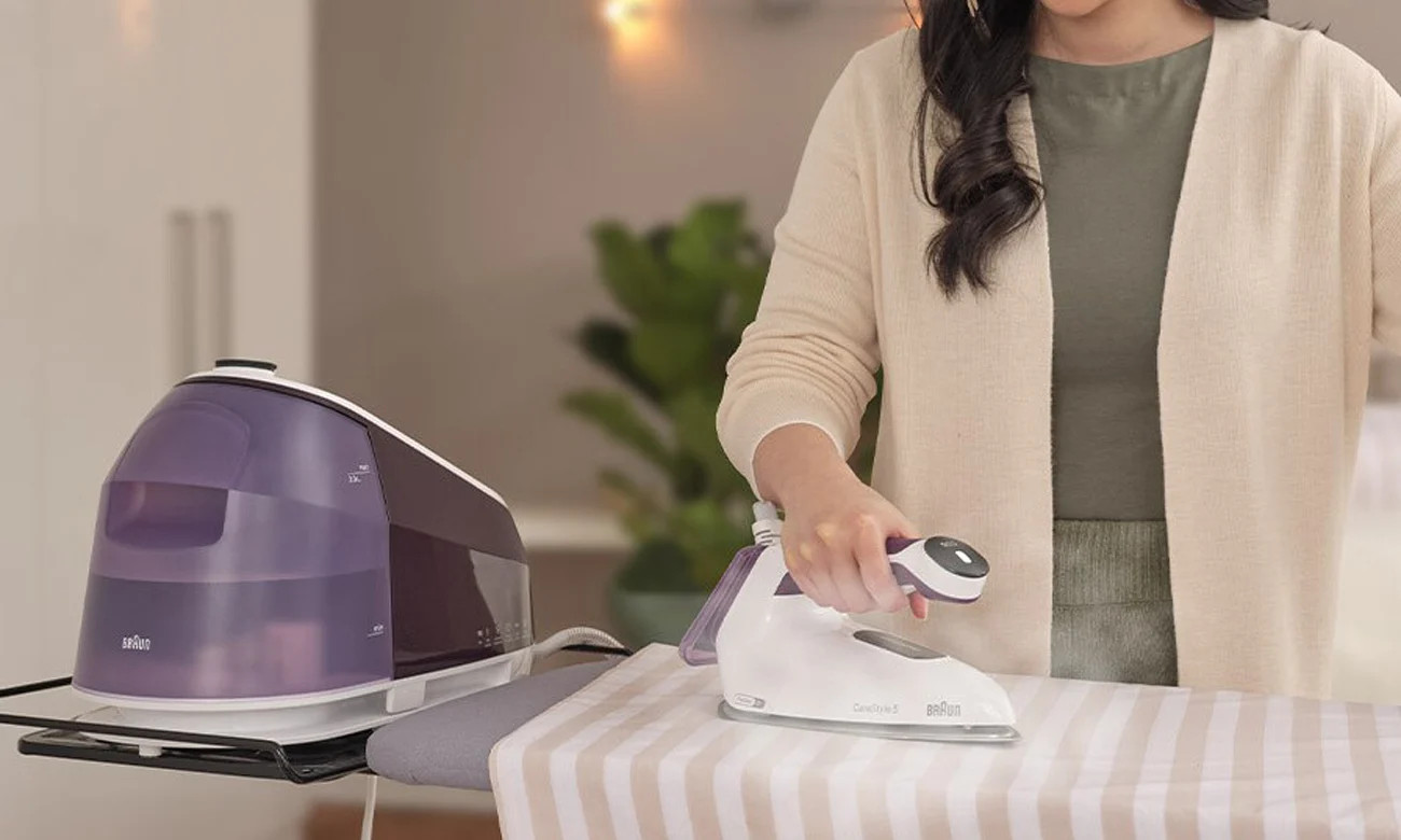 Kobieta prasująca pasiastą tkaninę na desce do prasowania, żelazko parowe i fioletowy zbiornik.