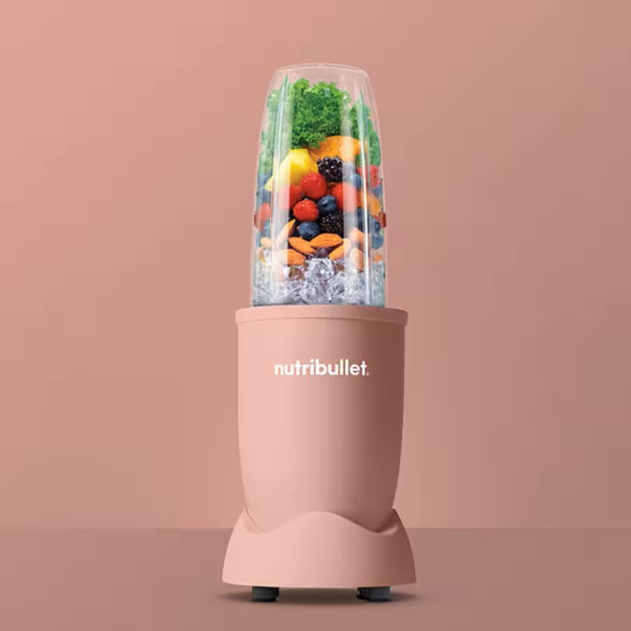 Różowy blender Nutribullet wypełniony owocami i lodem, na różowej powierzchni.