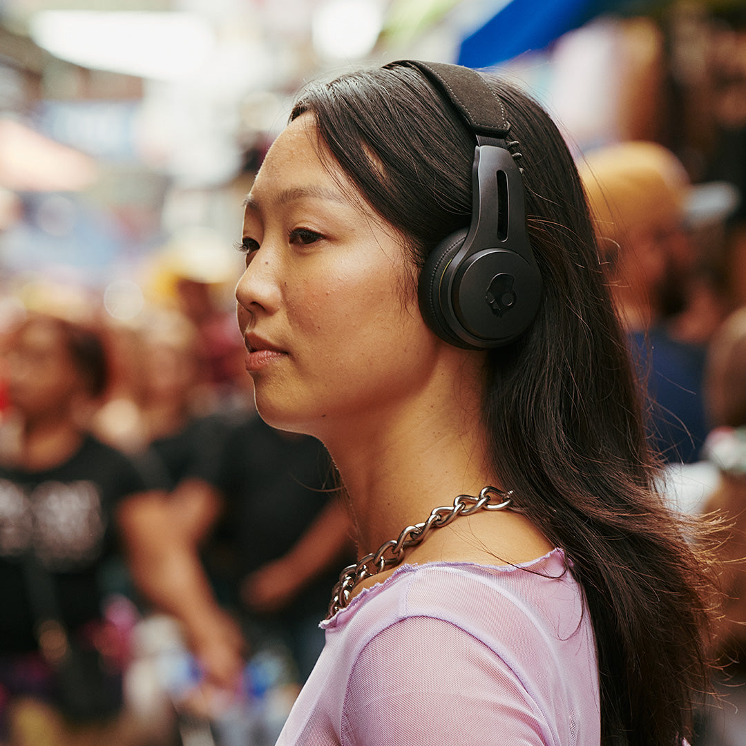 Eine Frau in rosa Hemd trägt schwarze Kopfhörer in einer belebten Straßenszene.