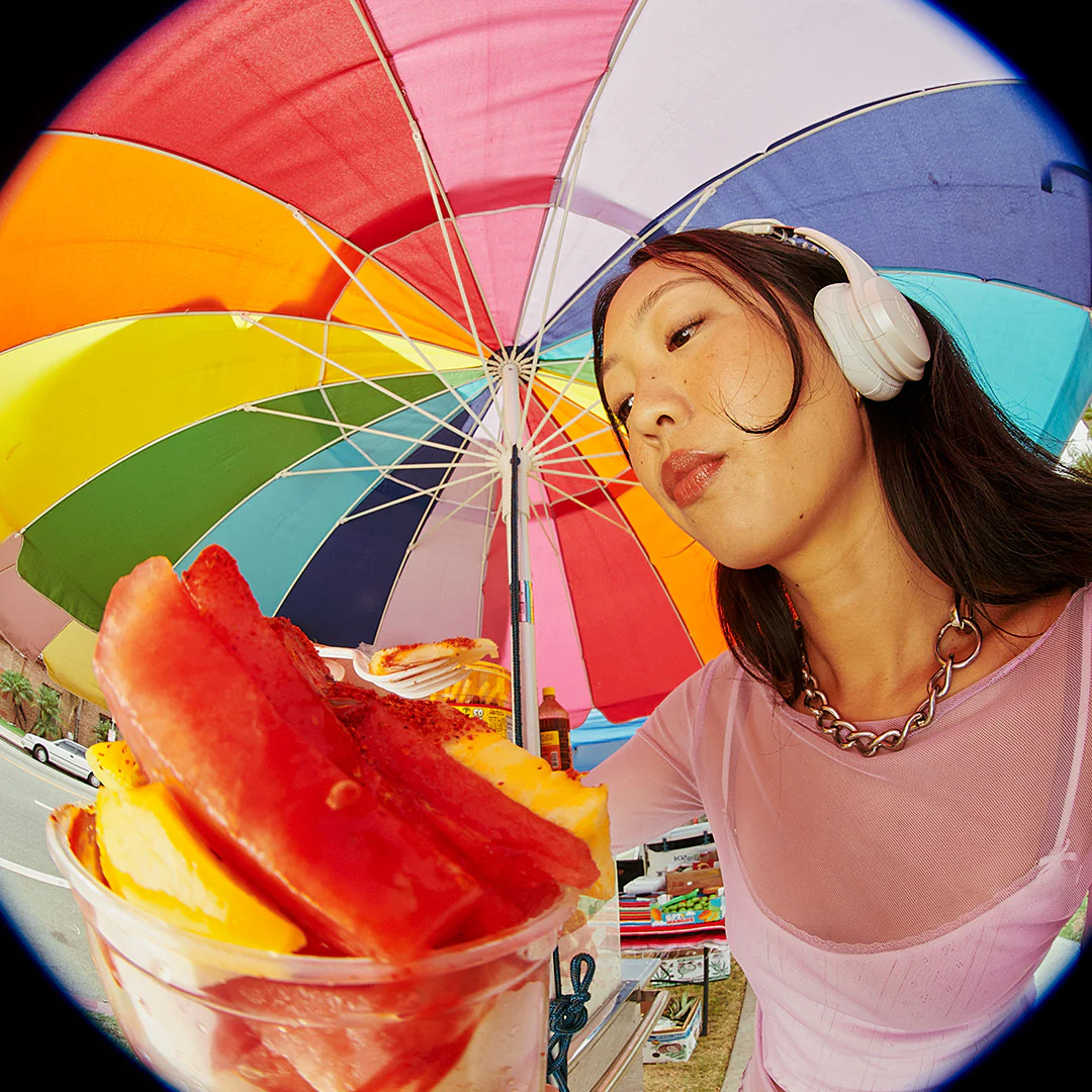 Frau mit Kopfhörern und einem bunten Regenschirm mit einer Tasse Snacks.