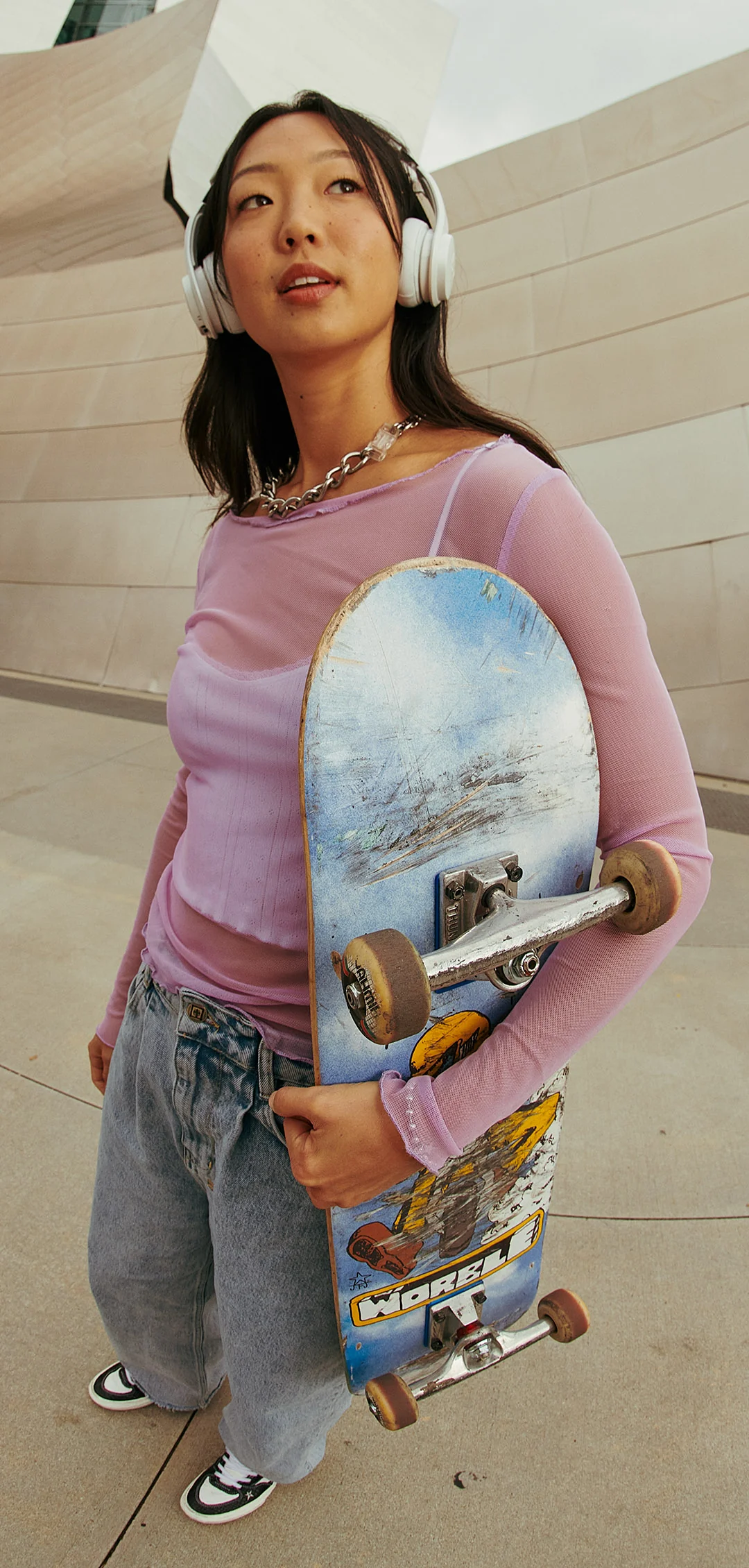 Junge Frau mit Kopfhörern und Skateboard. Sie trägt ein rosa Hemd und Jeans.