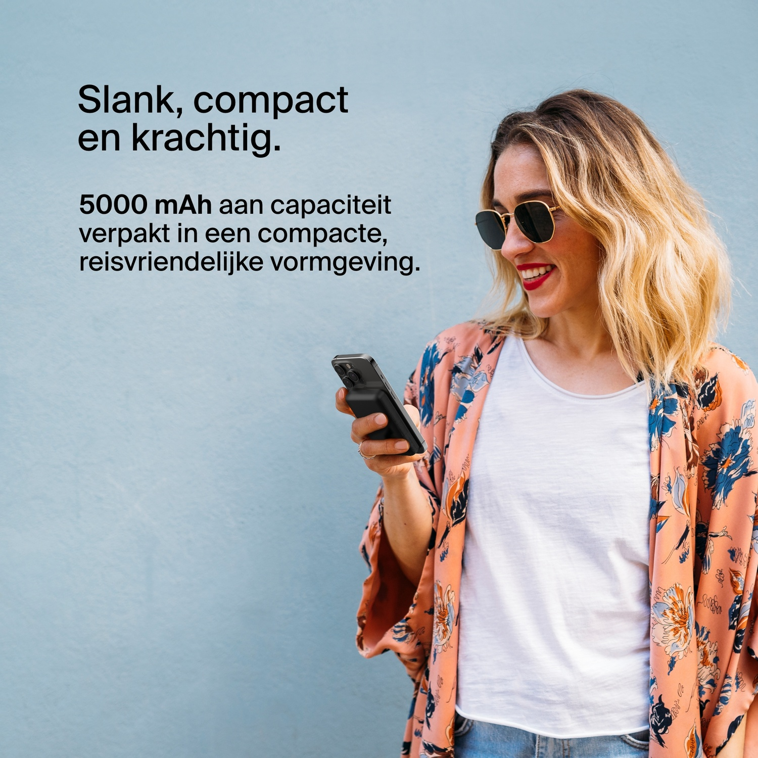 Vrouw met zonnebril houdt telefoon vast, draagt ​​een bloemen top, blauwe achtergrond.