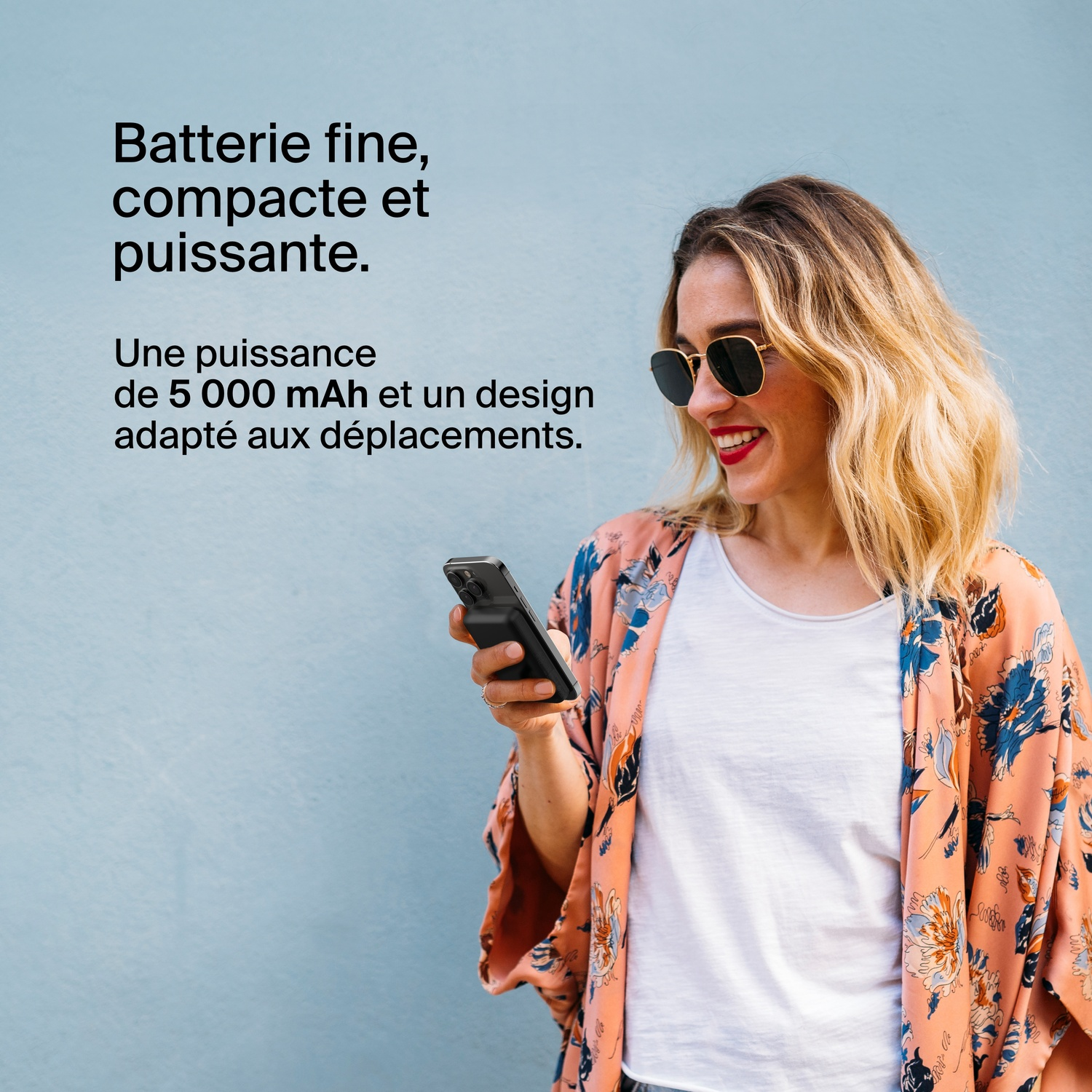 Une femme tenant un téléphone avec une batterie externe, portant une veste colorée et des lunettes de soleil.