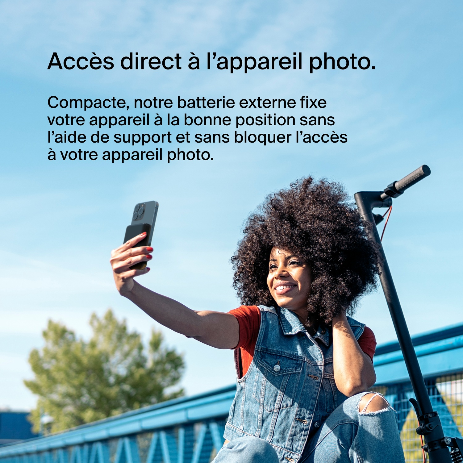 Une femme prend un selfie avec un smartphone fixé à un appareil noir. Un pont bleu et des arbres en arrière-plan.