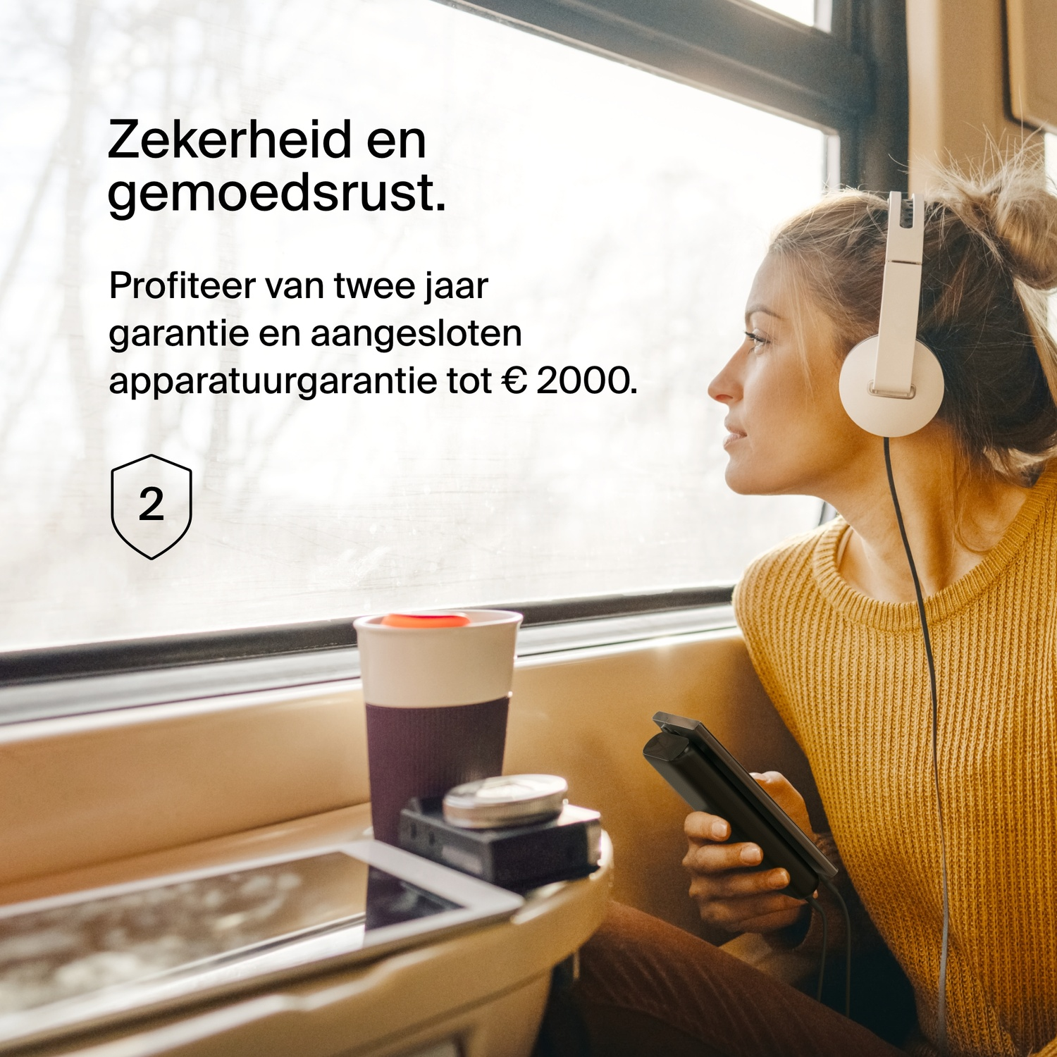 A person looking out a train window, wearing headphones. A phone and a cup are nearby.