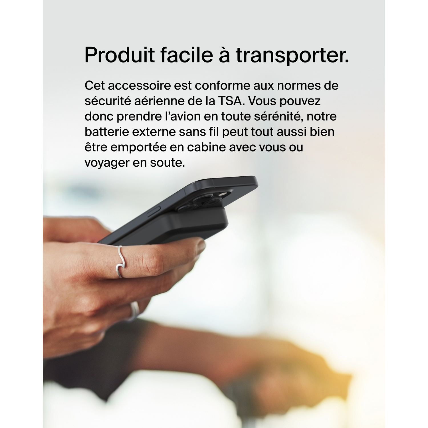 Mains tenant un téléphone noir avec un accessoire noir. Une personne est floue en arrière-plan avec la lumière du soleil.